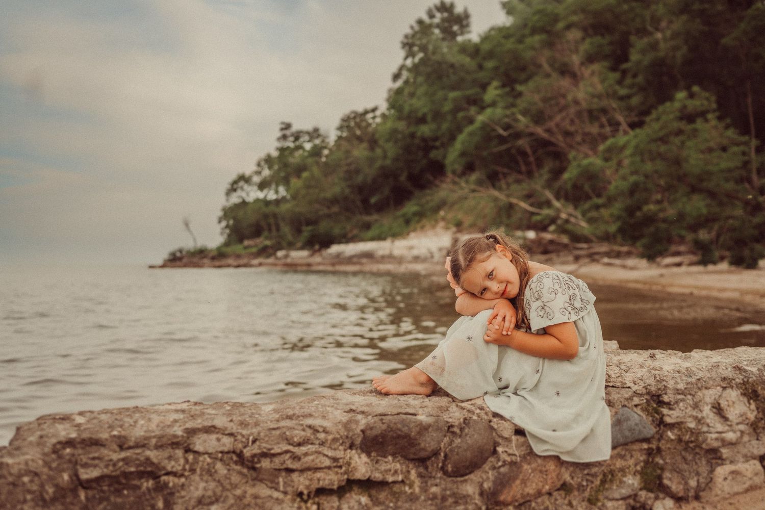 Child in light dress explores rocky coastline during golden hour.