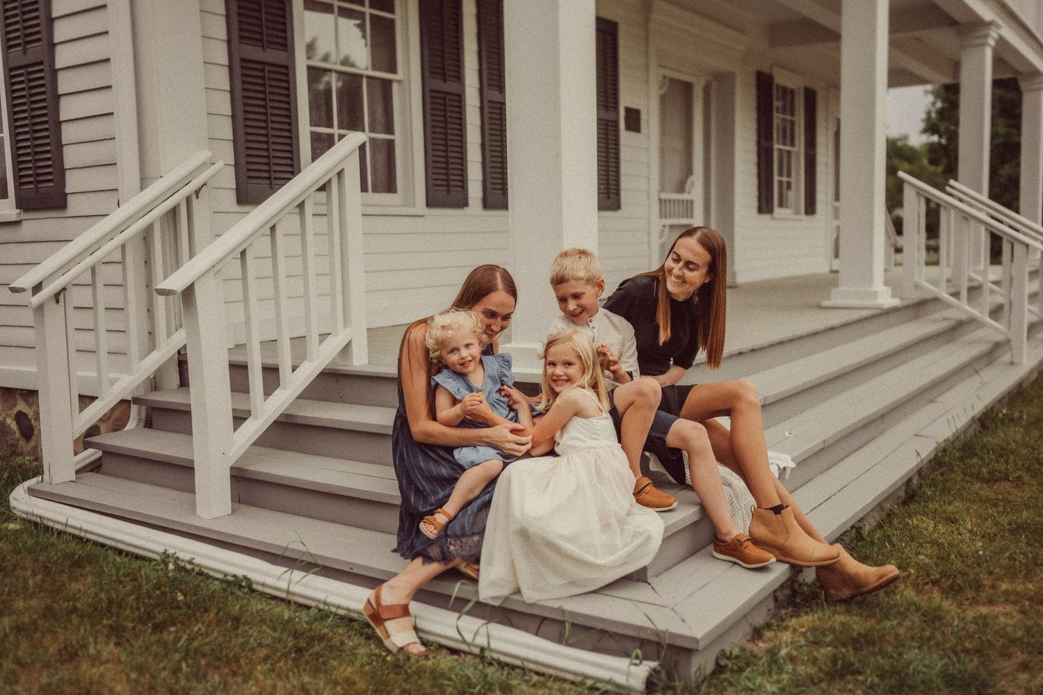 Group sitting casually on white porch steps of classic colonial house on summer day.