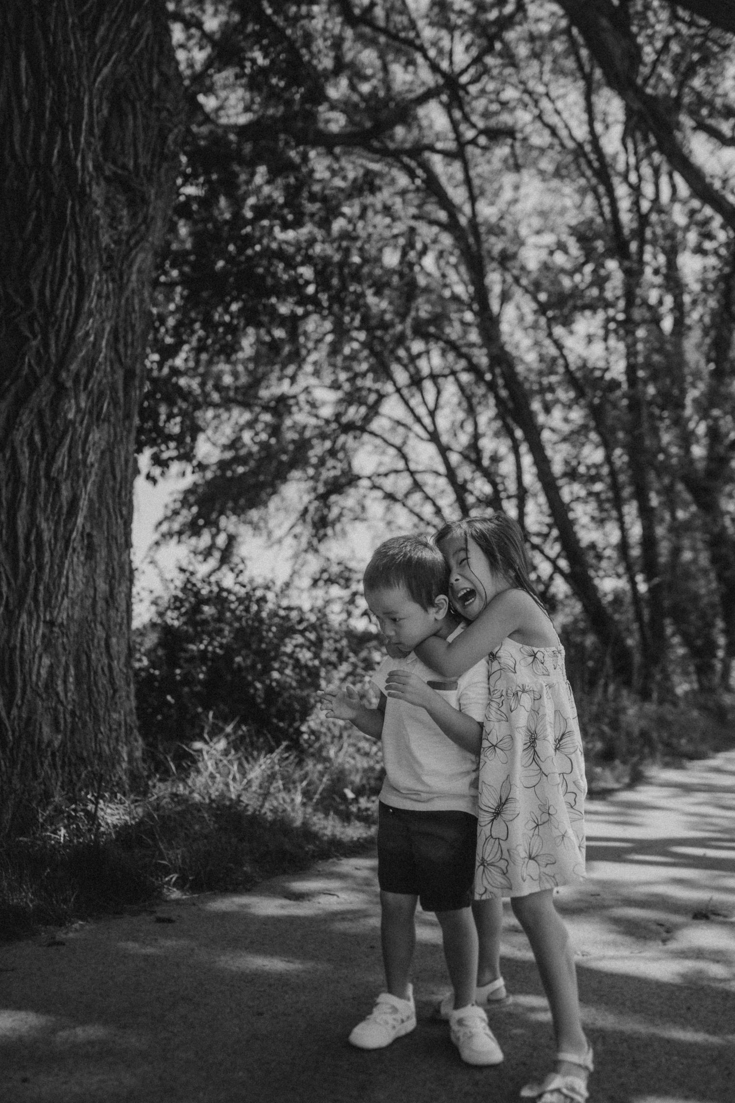 Two small children share a sweet moment on a tree-lined path in black and white.