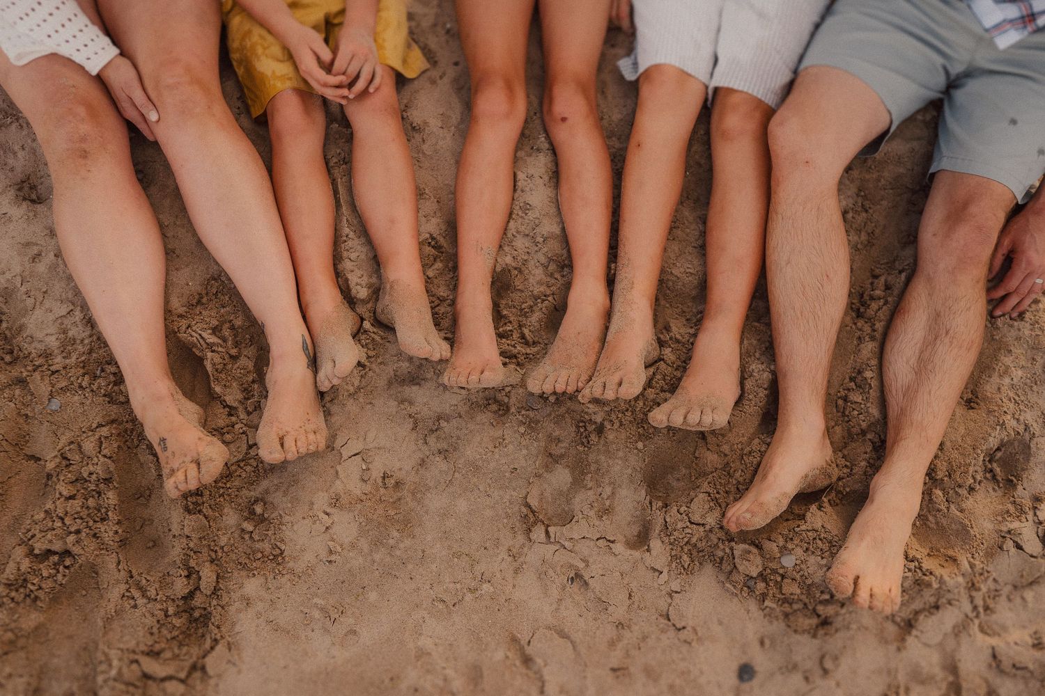 Row of bare feet and legs resting on sandy beach surface during summer leisure activity.