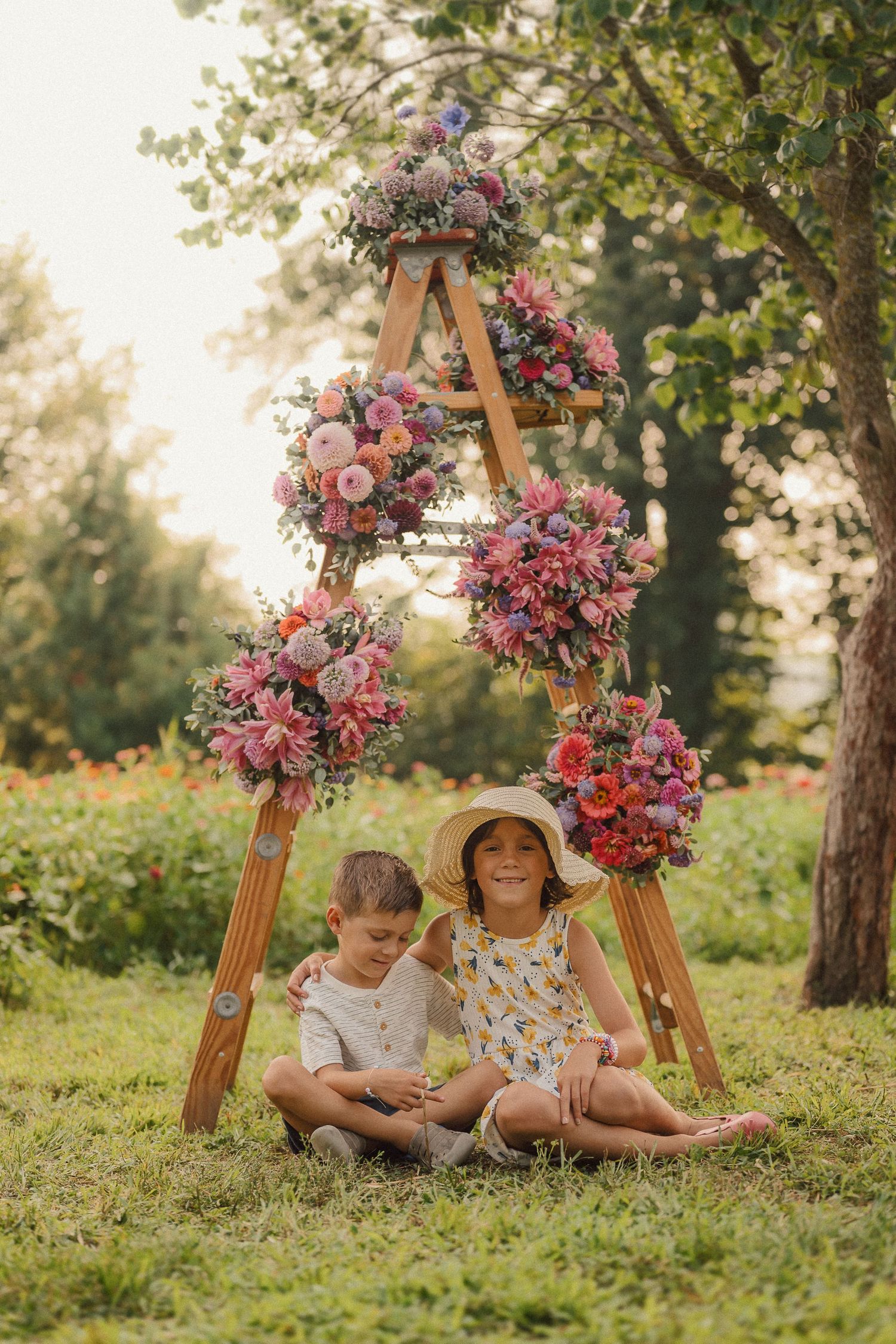 Two children sitting under a wooden triangle ladder decorated with pink and purple floral arrangements in a garden setting.