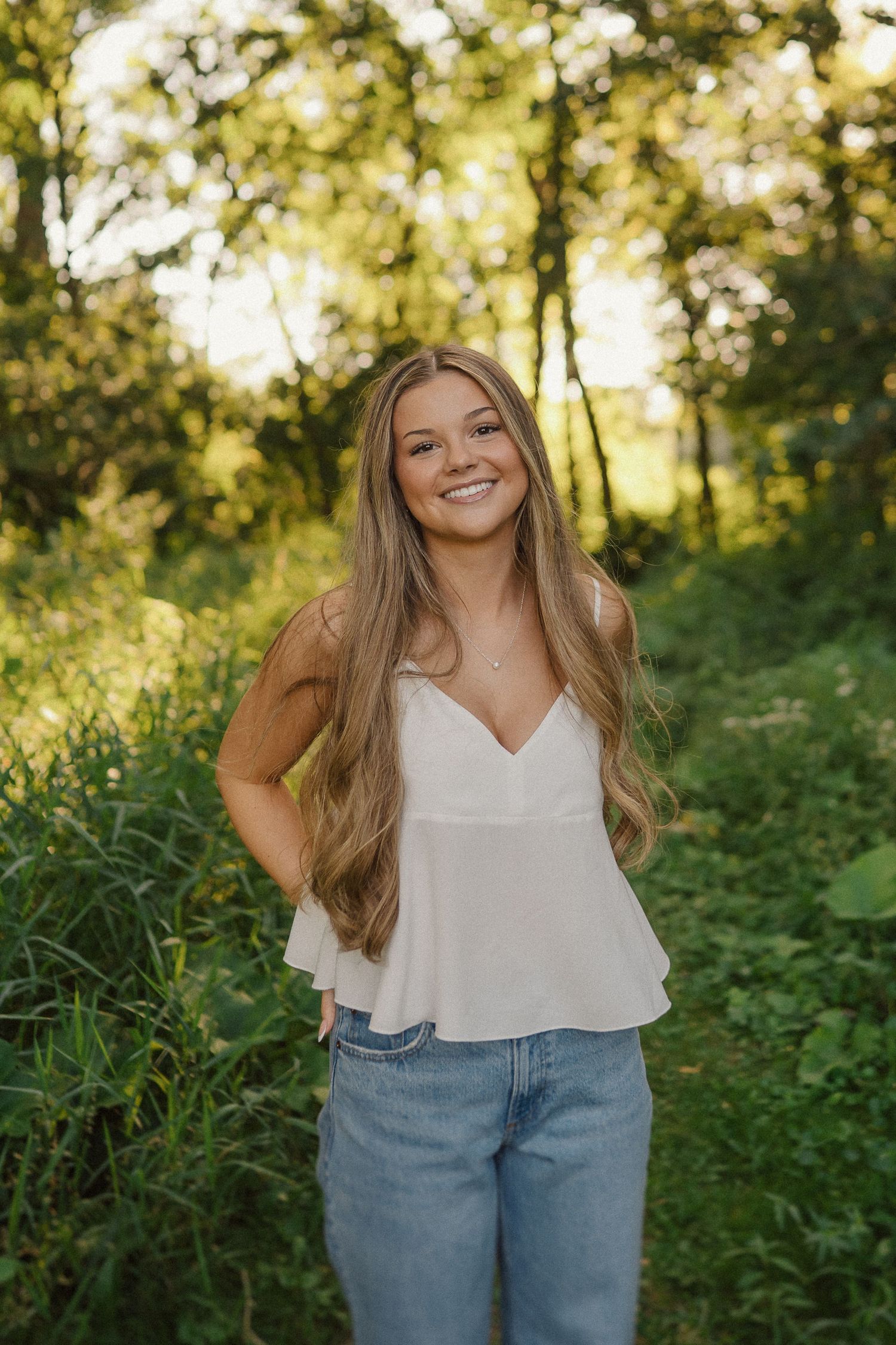 A woman wearing a white camisole and jeans smiles brightly while standing in a lush green forest setting.