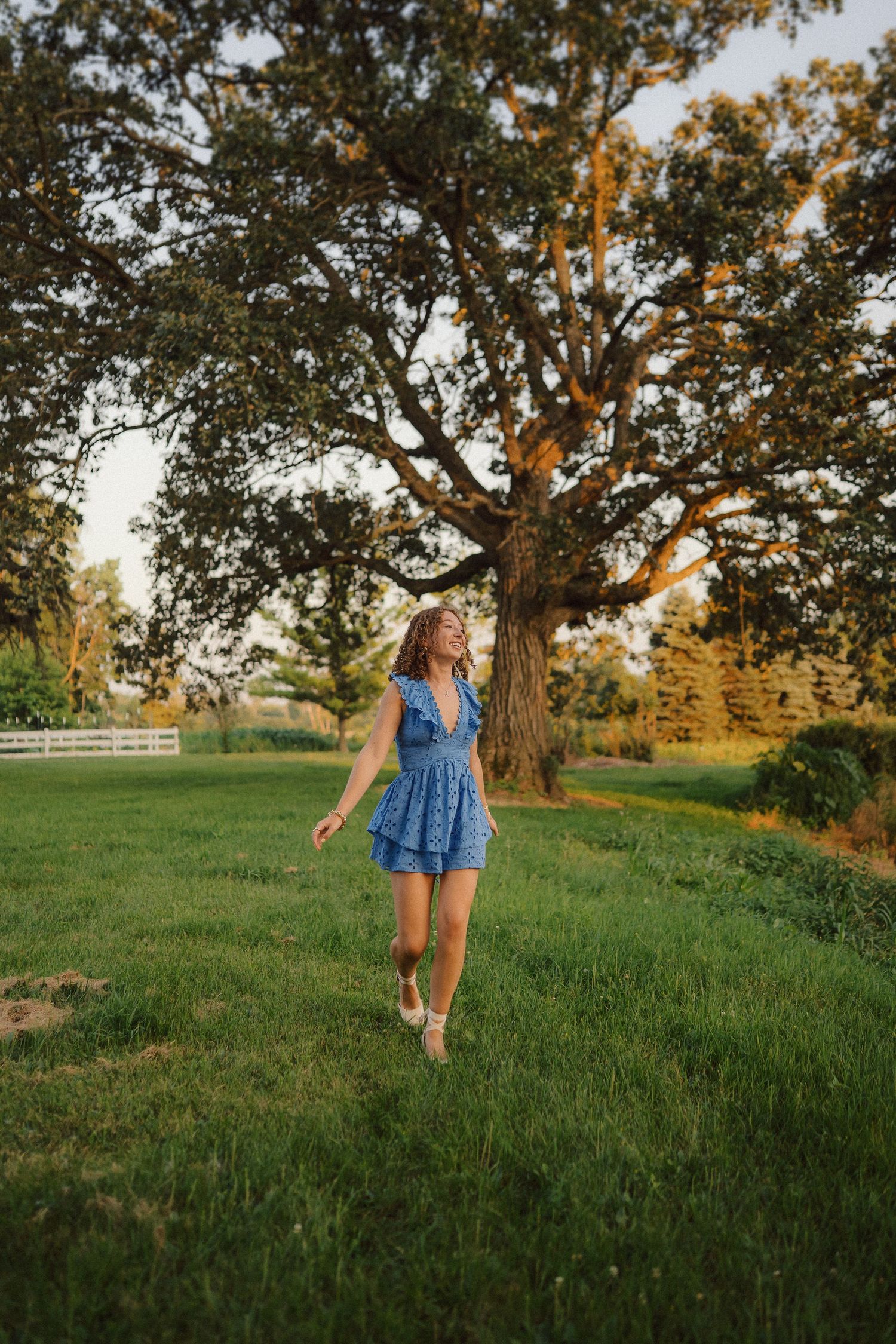 A person in a blue dress twirls joyfully under a large oak tree during golden hour in a grassy field.