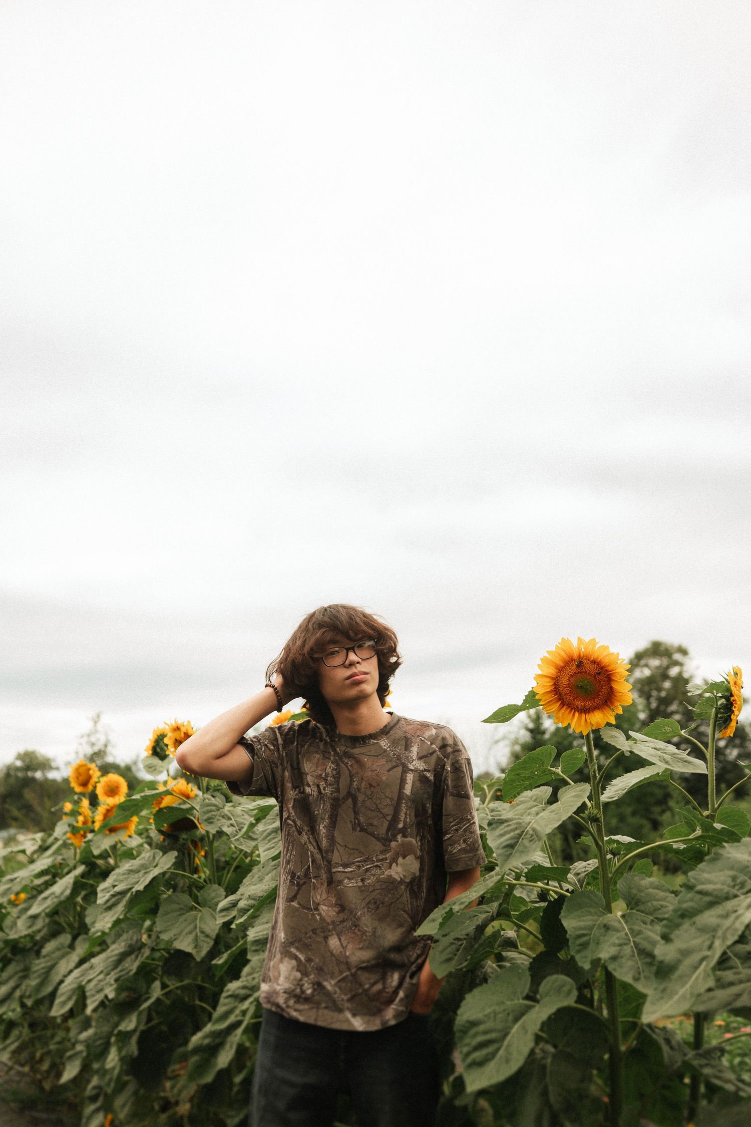 A person stands in a field of tall sunflowers and green foliage under a cloudy sky.