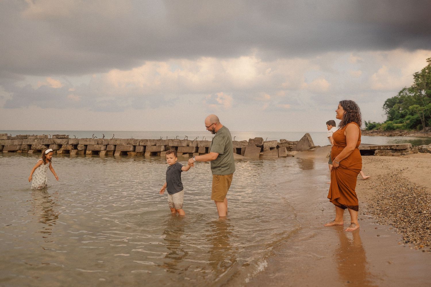 Family members wade in shallow water near a rocky shoreline during sunset.