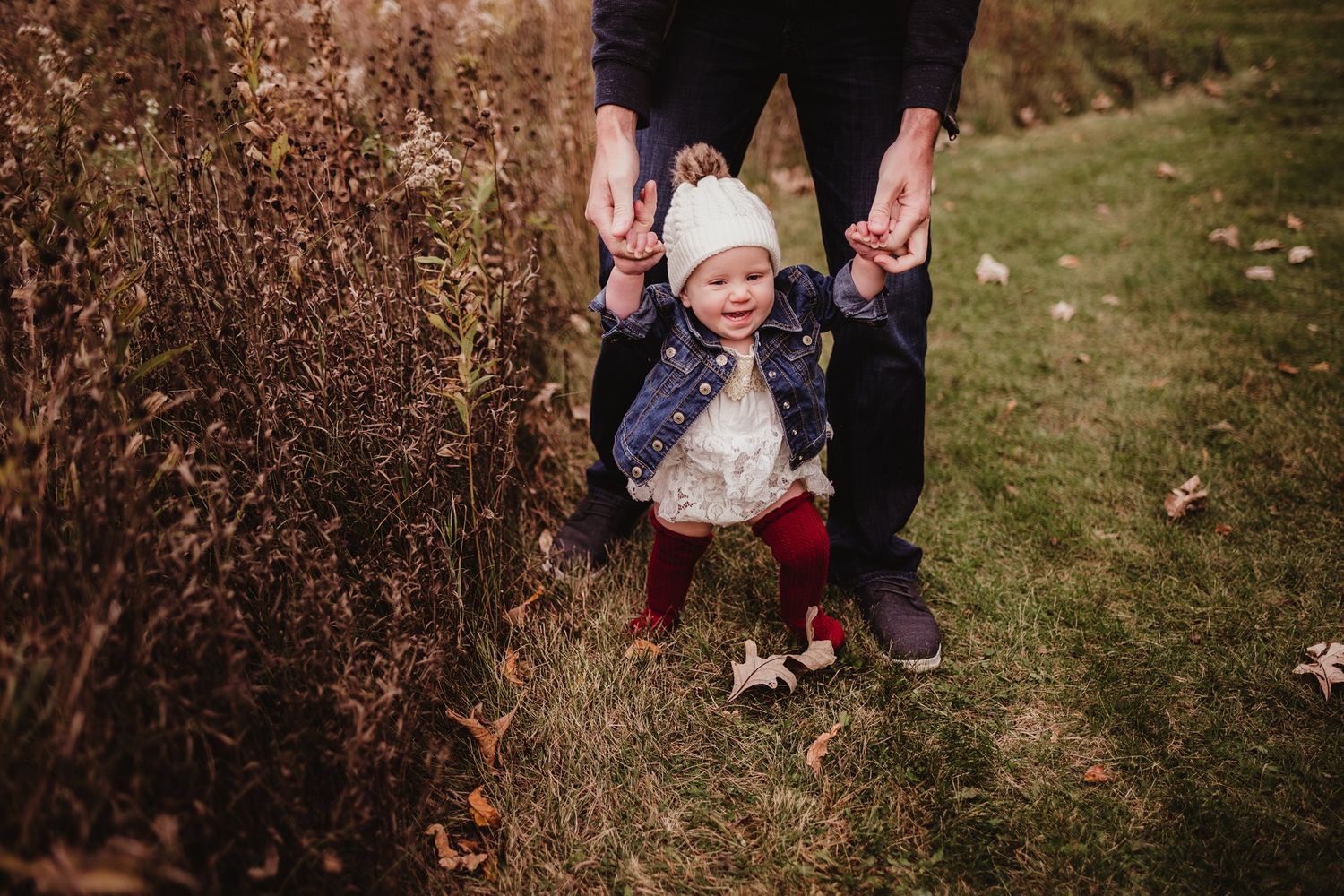 Toddler walking between adult hands on autumn path surrounded by dried plants.