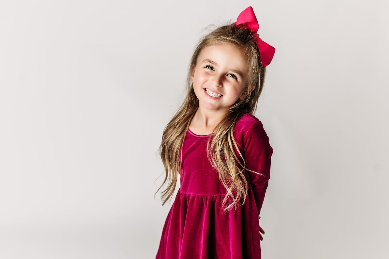 A happy child wearing a velvet burgundy dress and pink bow hair accessory poses against a white backdrop.