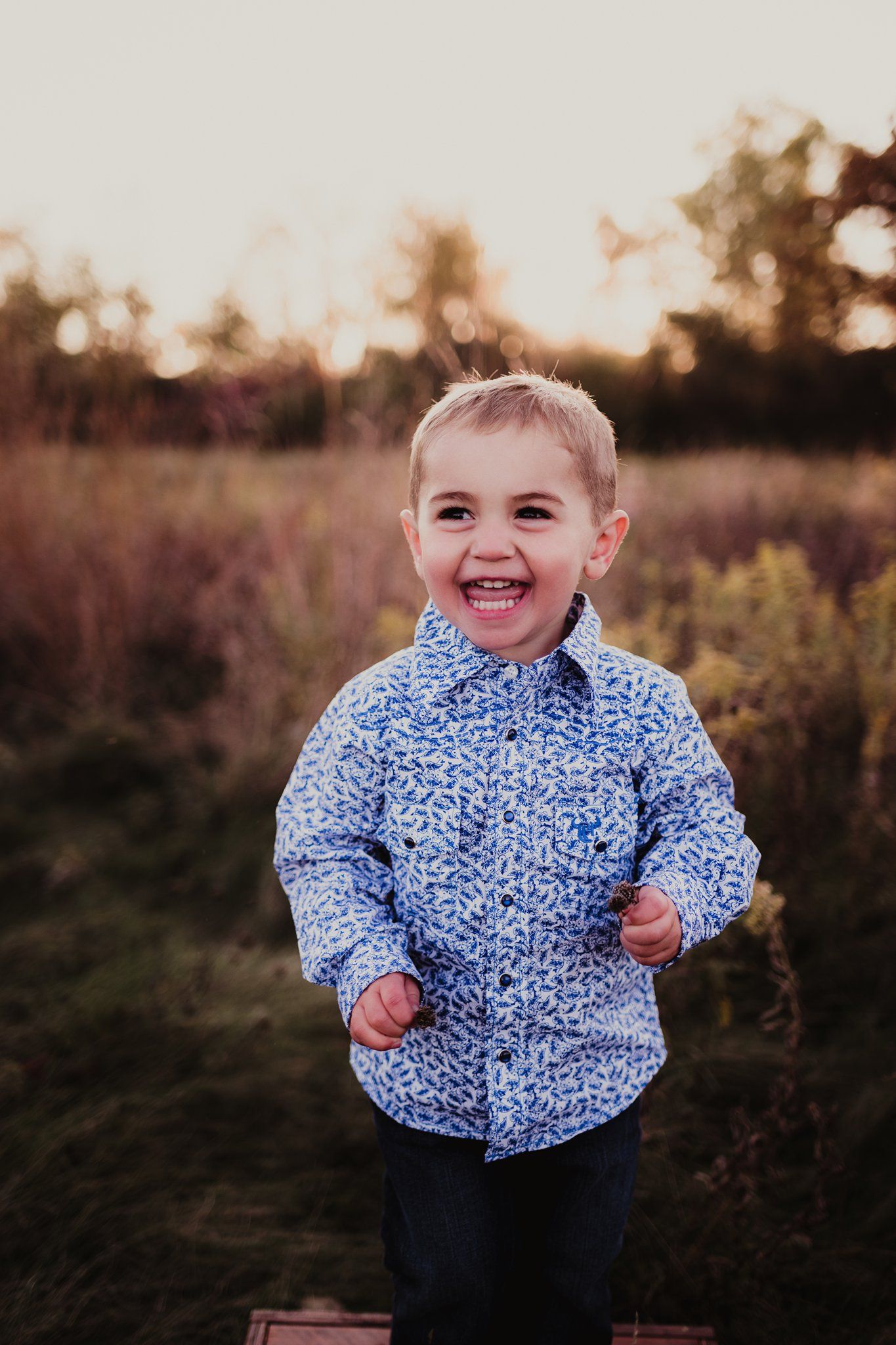 A laughing child wearing a blue patterned shirt outdoors at sunset.