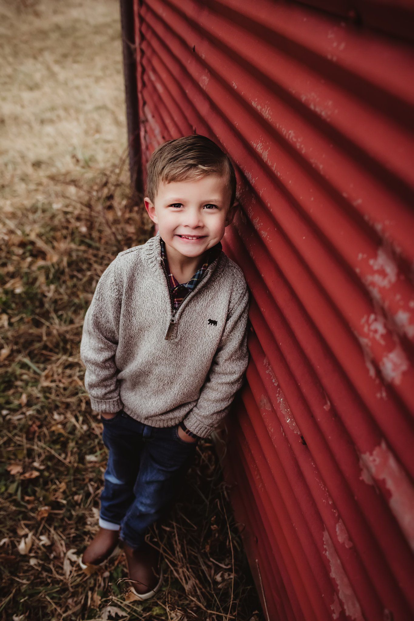 A child in a grey sweater poses against a red corrugated metal wall with a bright smile.