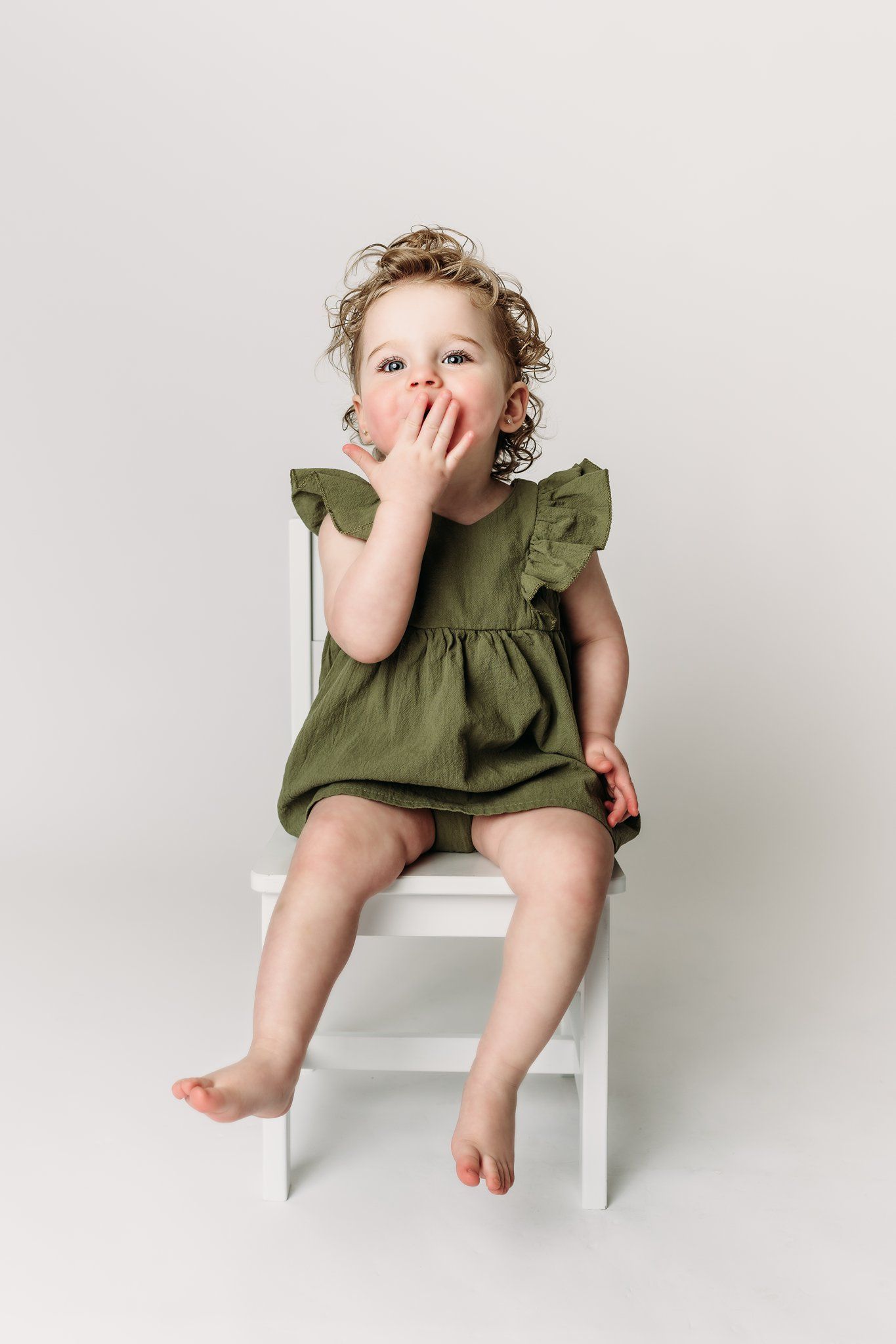 Child in olive dress sits on chair with hand covering mouth playfully.