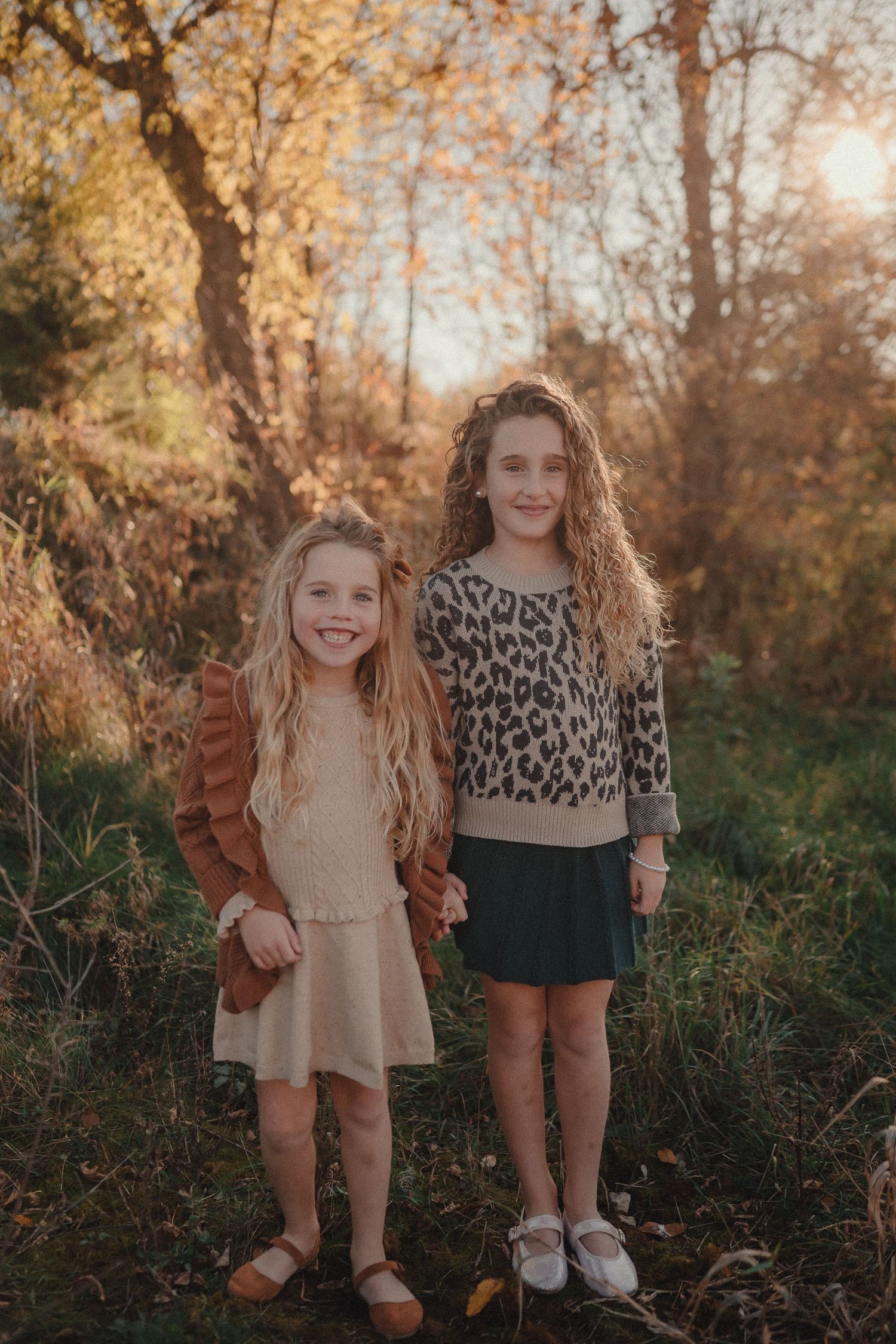 Two children pose together in a sunlit autumn forest wearing casual clothing.