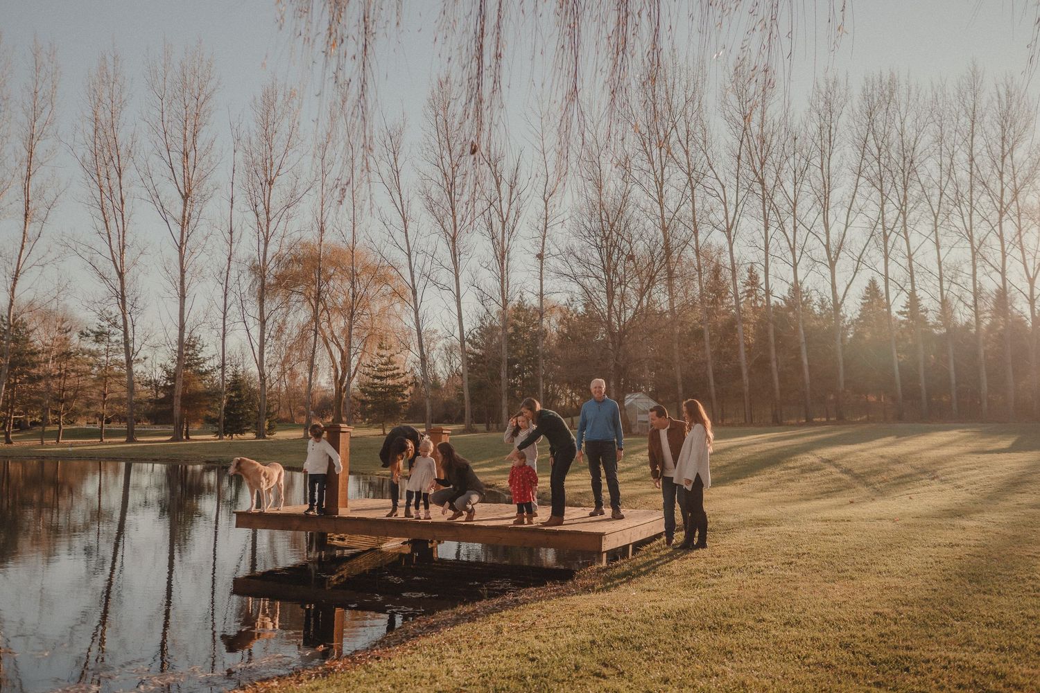 A group gathers on a wooden dock by a pond during sunset with bare trees in the background.