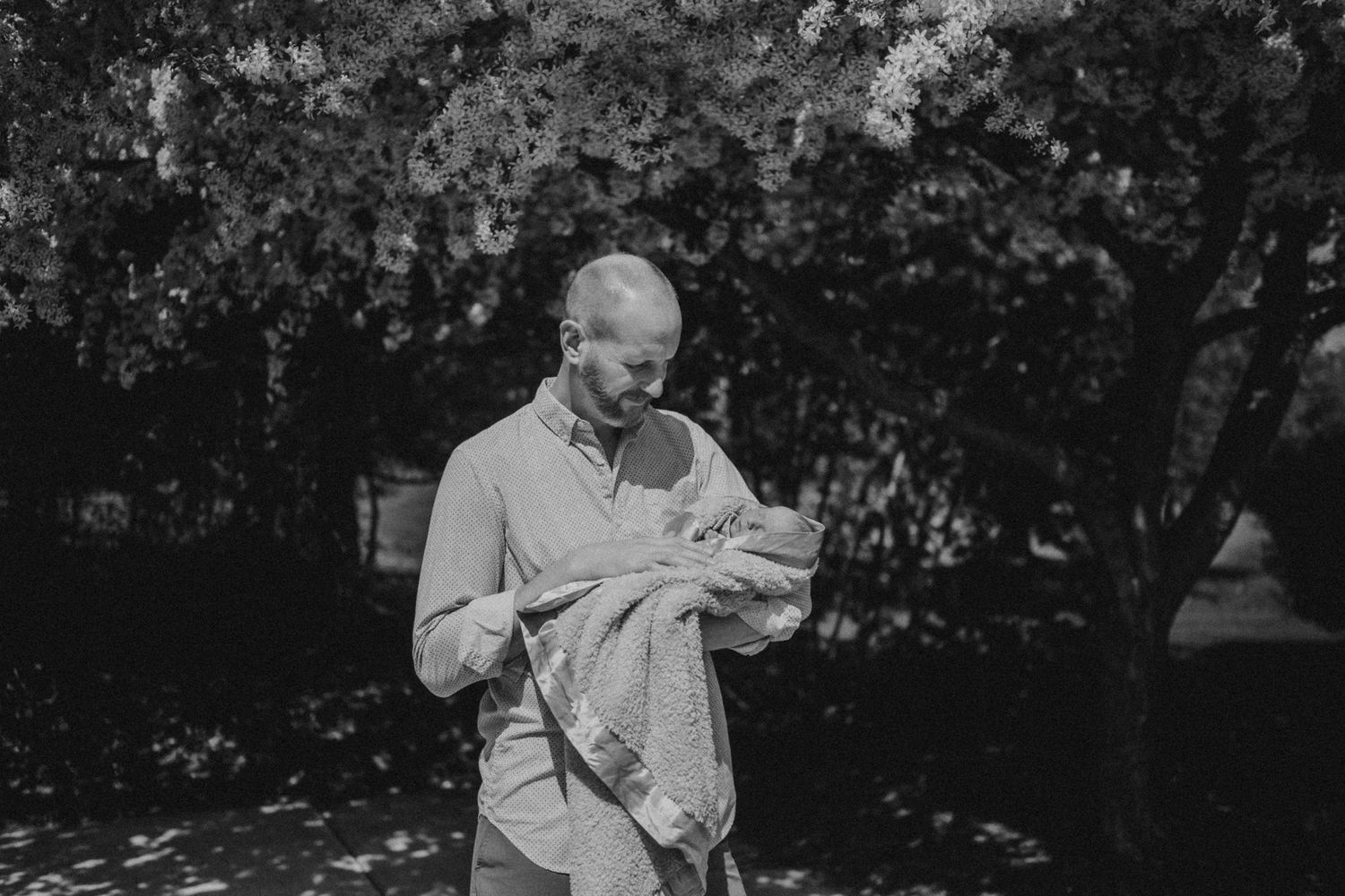 Black and white portrait shows someone cradling a newborn baby outdoors under trees.