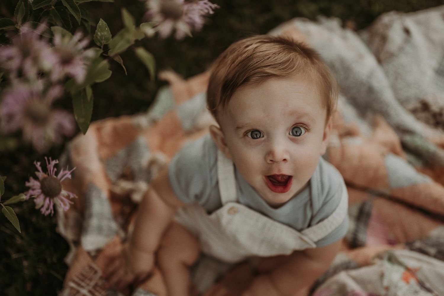Joyful baby in gray outfit sits on patterned blanket with pink wildflowers in background.