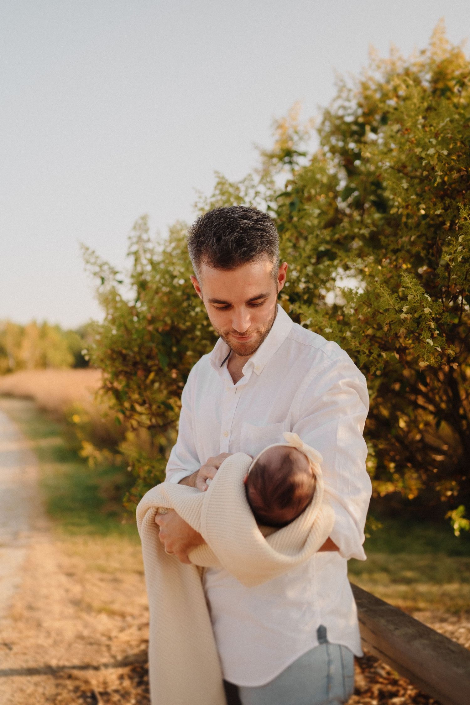 Outdoor portrait showing a person in white holding a swaddled infant near autumn trees on a country path.