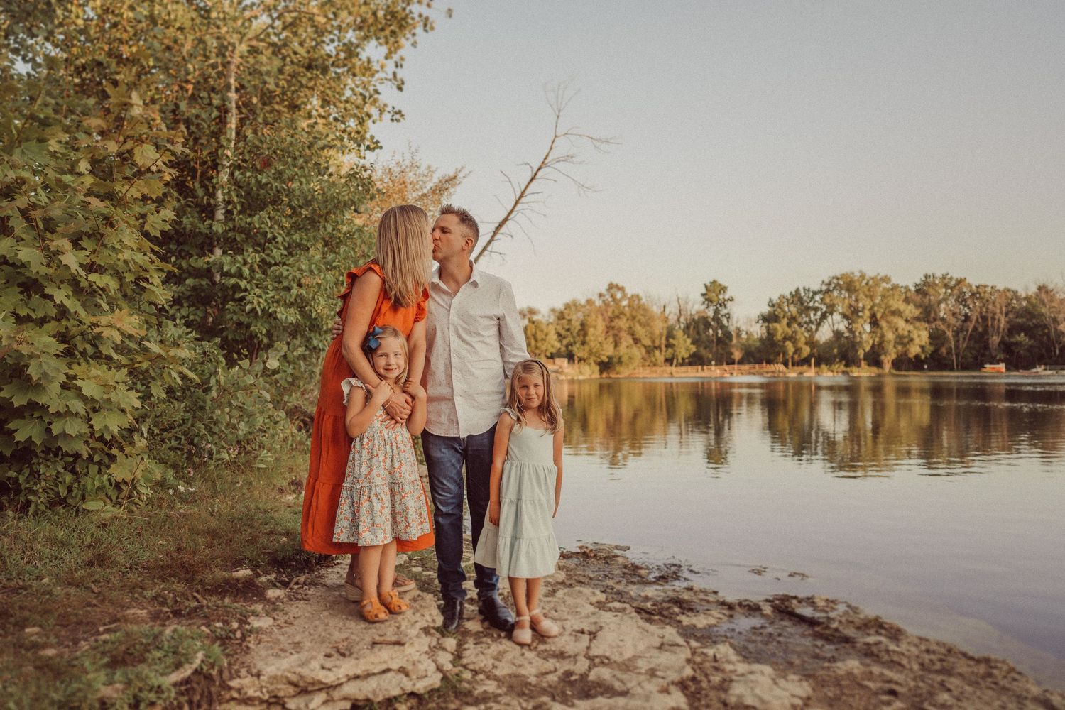 A family stands together on a rocky shoreline at sunset beside a tranquil lake surrounded by autumn trees.