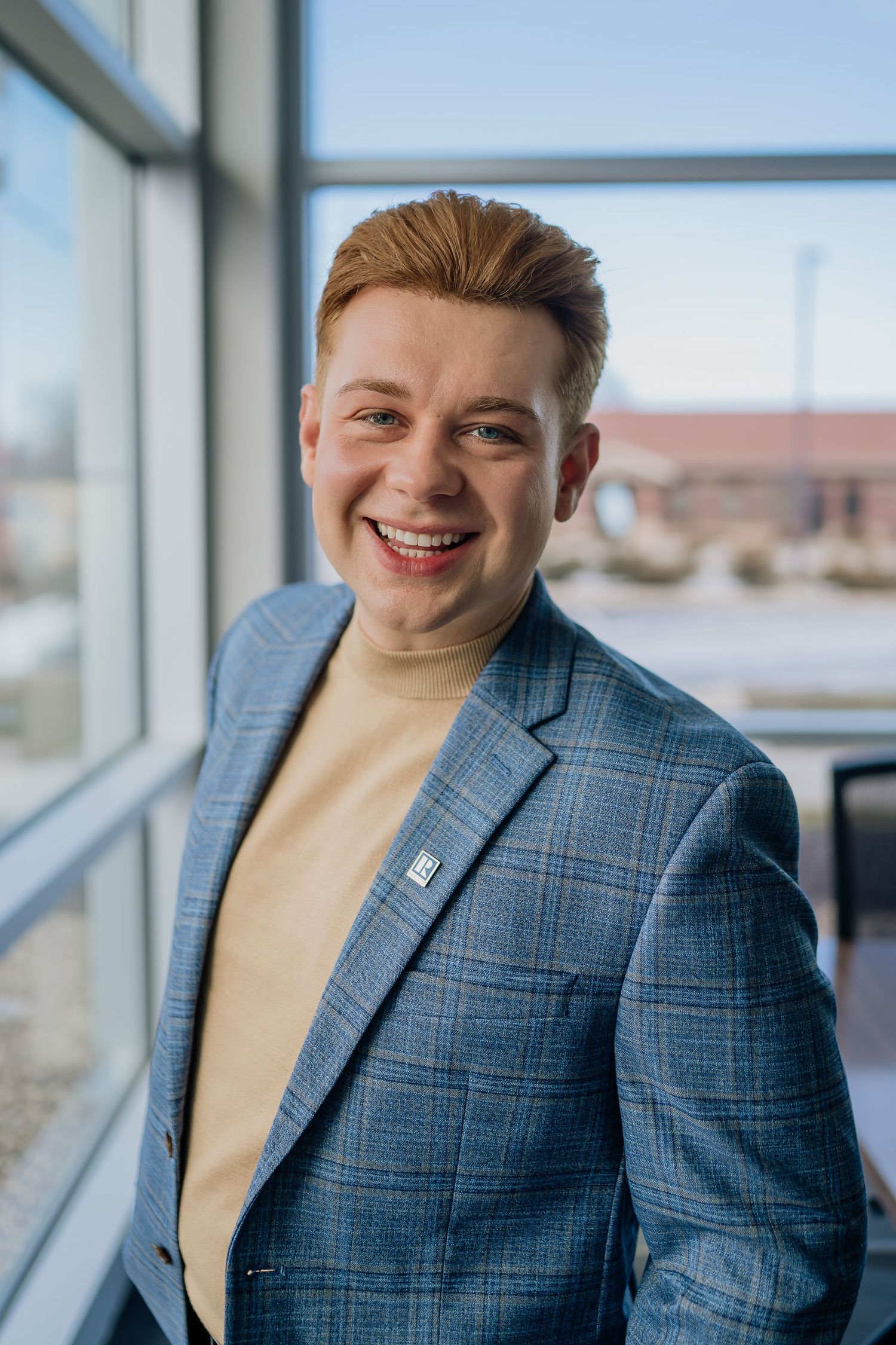 Person in stylish blue plaid blazer and beige turtleneck smiling near office windows with natural lighting.