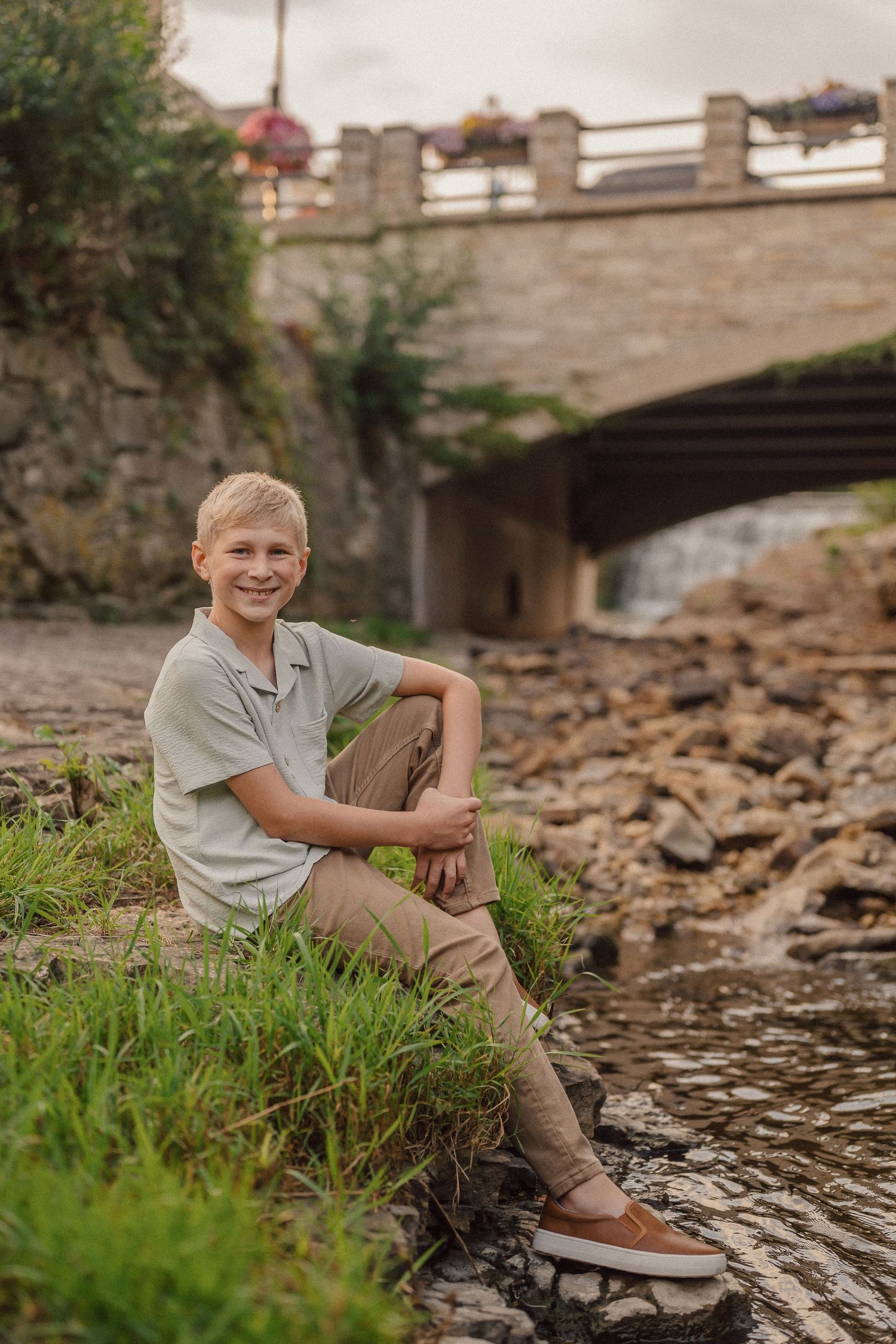 A young person sits casually on a rocky riverbank near a stone bridge on a sunny day.