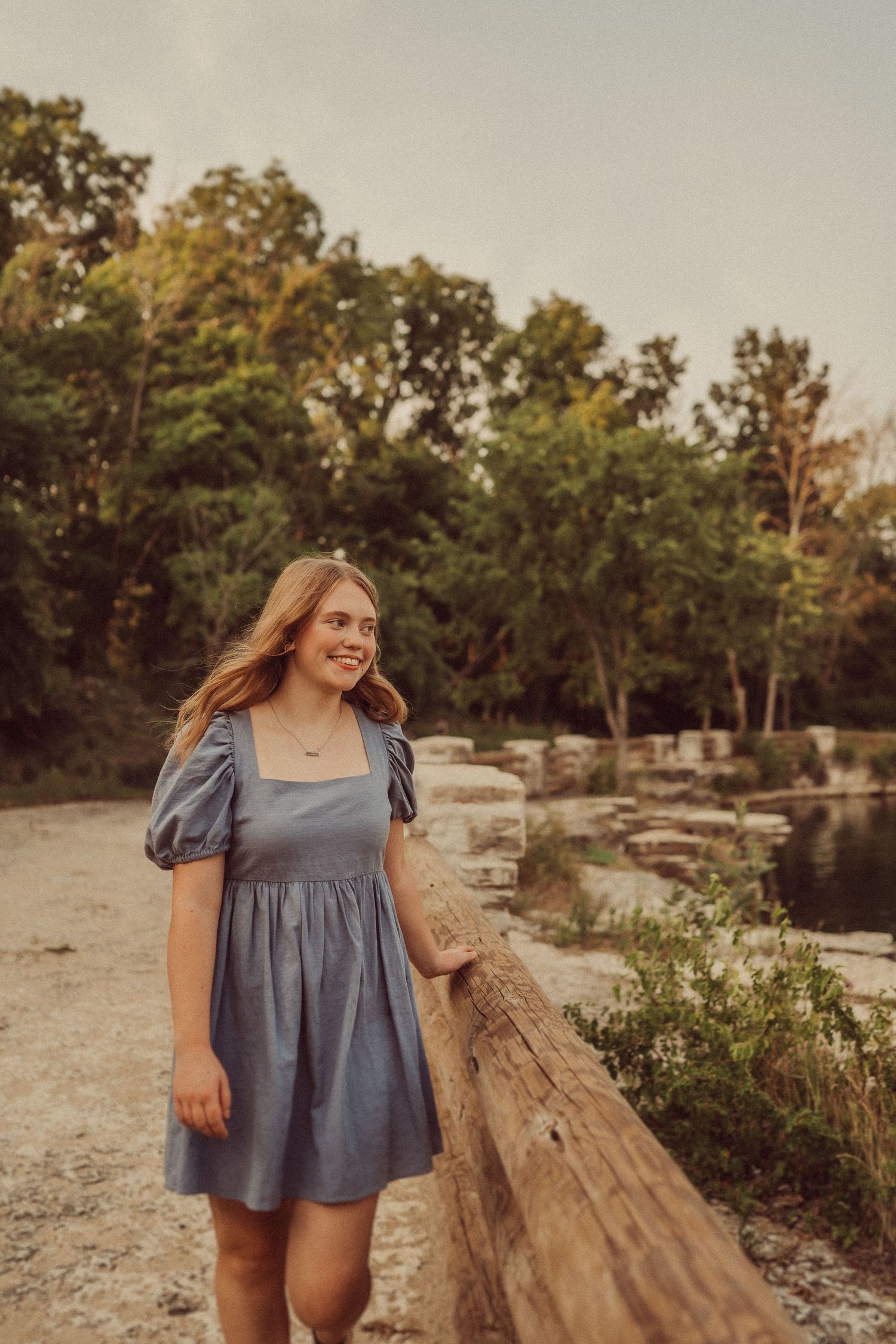 A blue dress-clad figure poses along a wooden railing near a scenic pond at sunset.