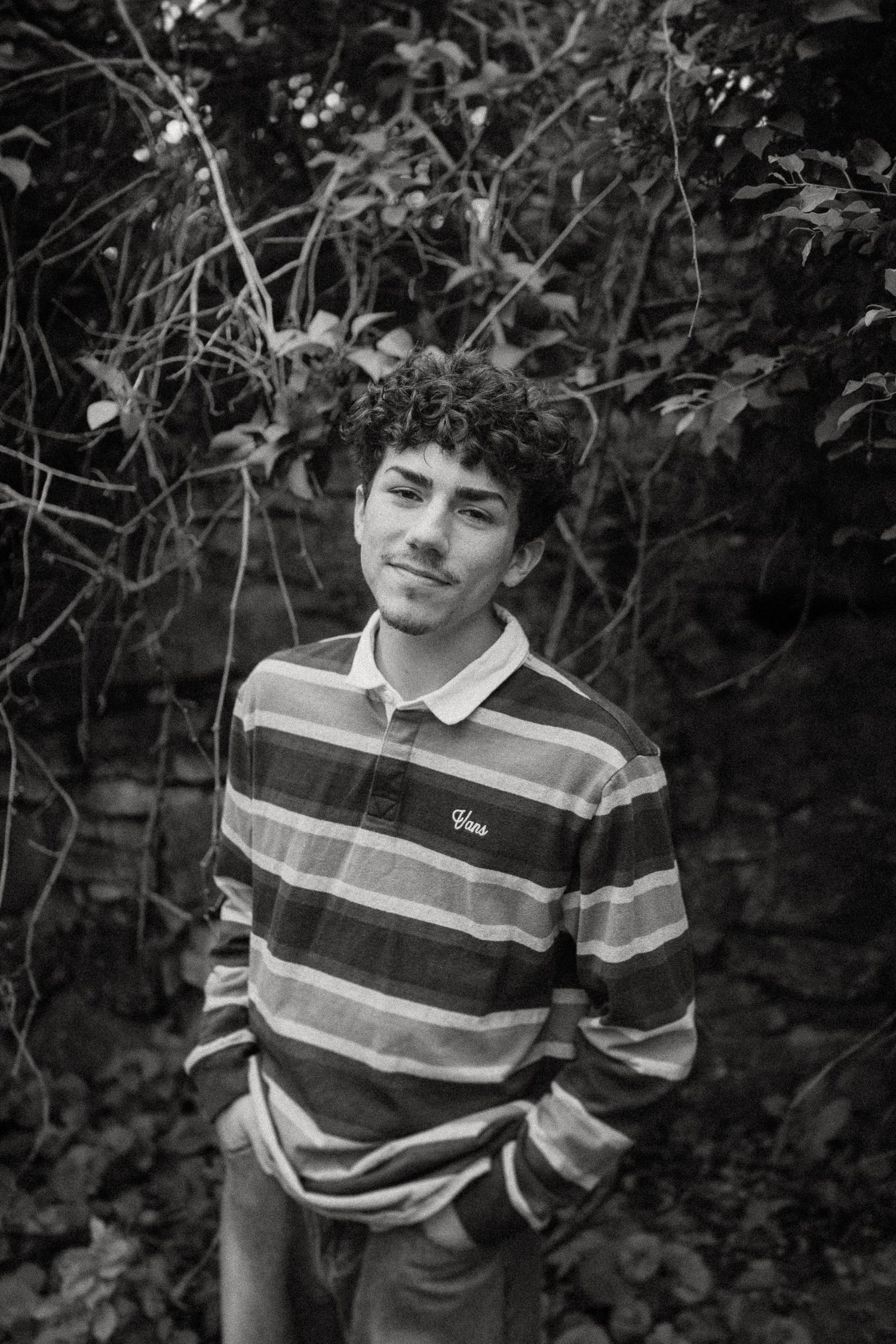 A black and white portrait of someone wearing a striped polo shirt against a natural foliage background.