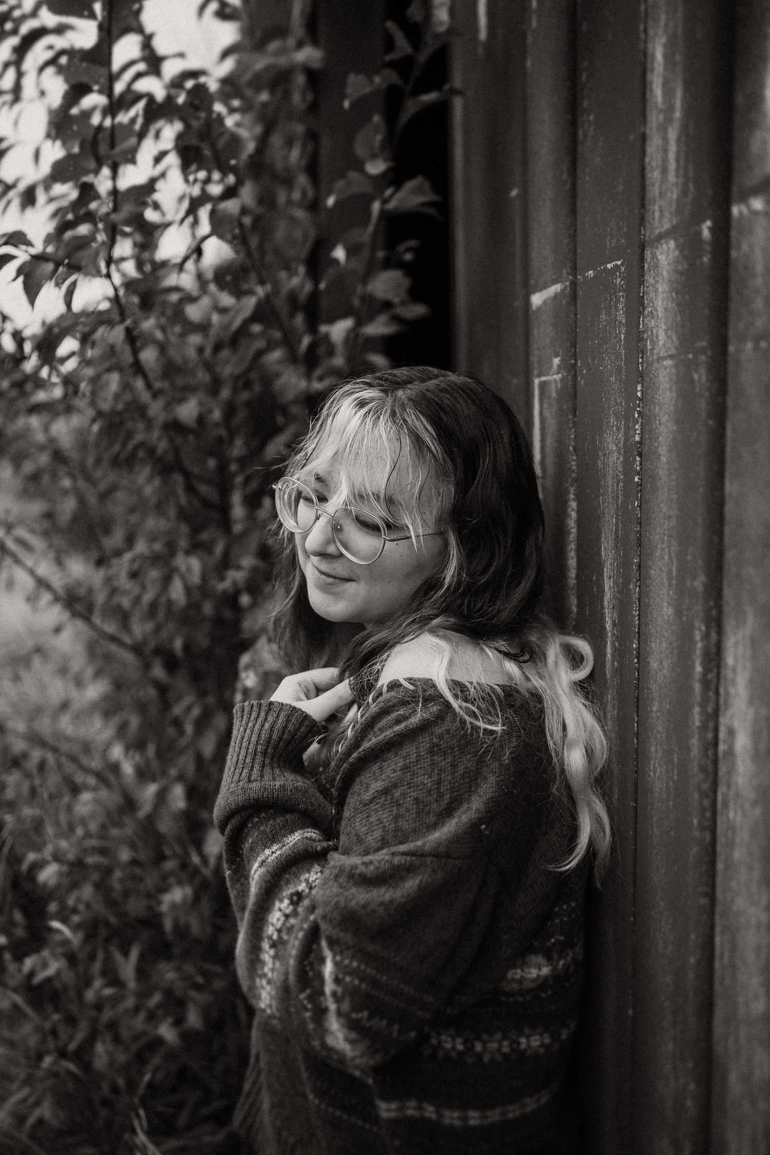A black and white portrait showing a joyful expression against rustic siding and foliage.