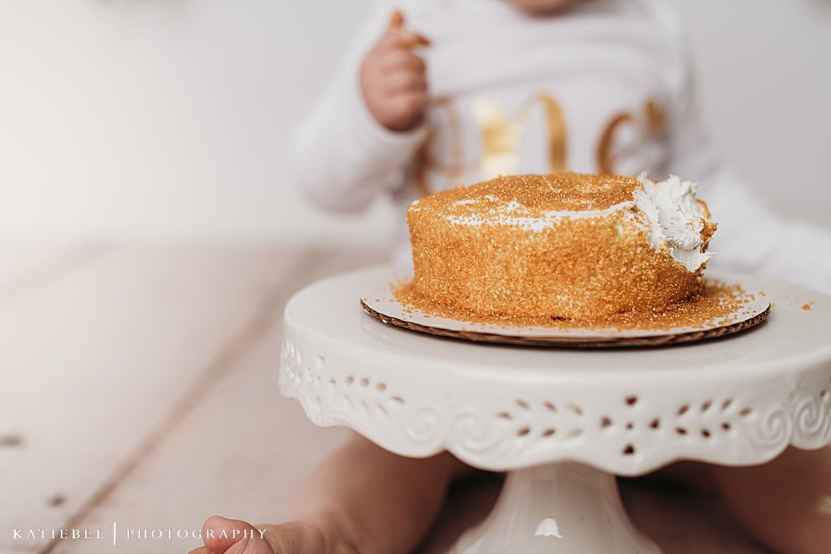 Close-up of golden birthday cake on white decorative cake stand with blurred baby in background.