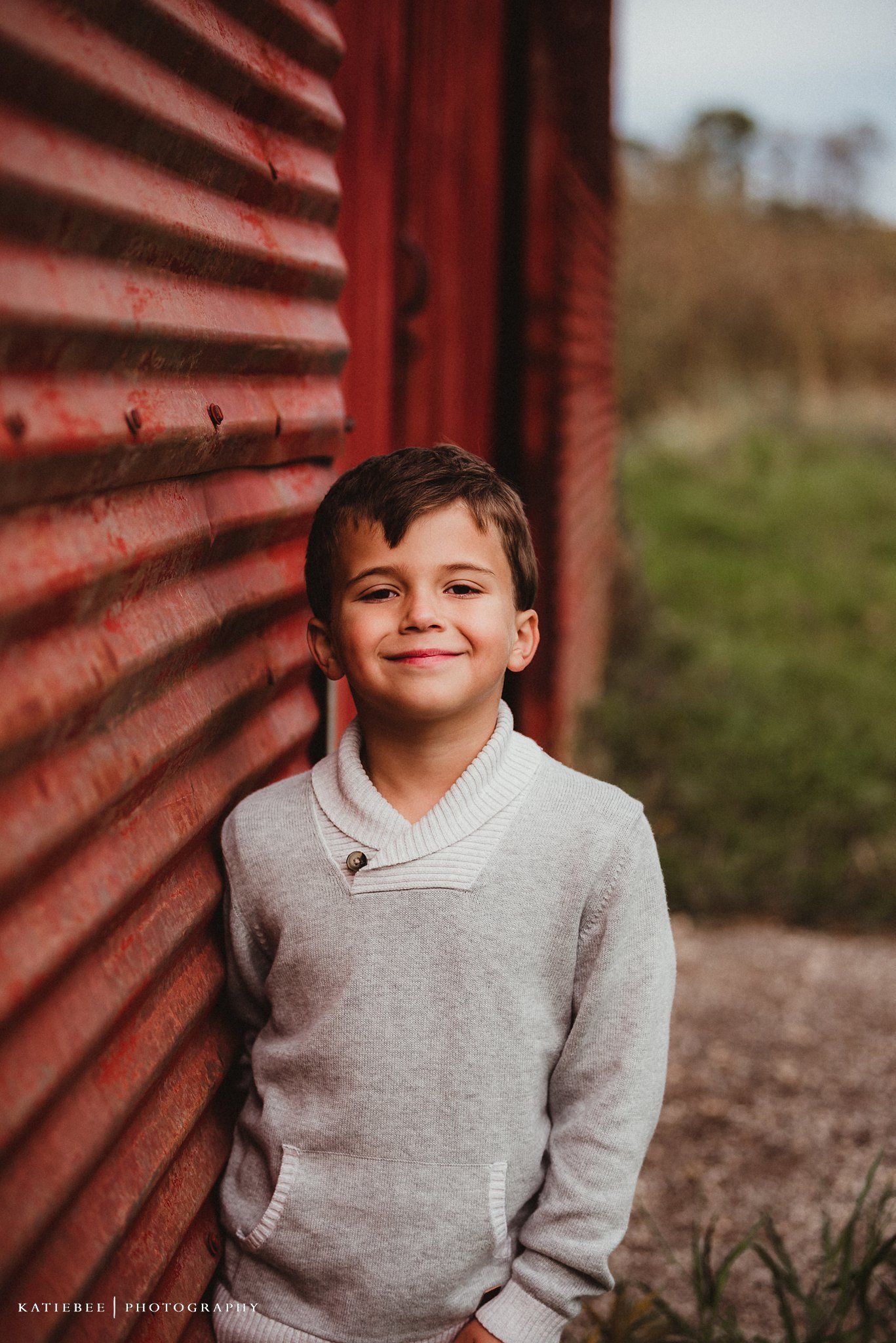 Portrait against red barn showing warm smile while wearing grey sweater.