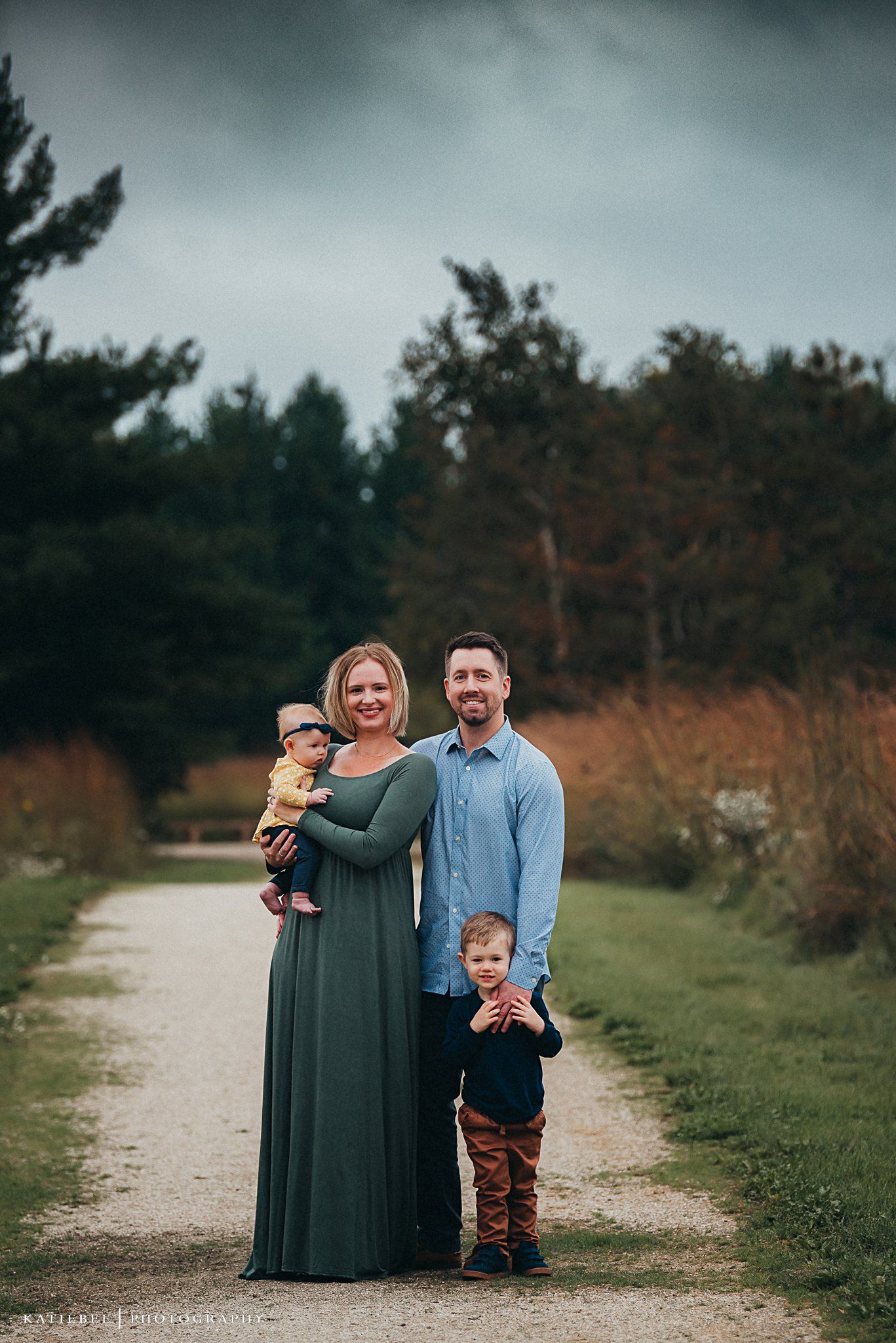 Family of four poses together on a path surrounded by evergreen trees on an overcast day.