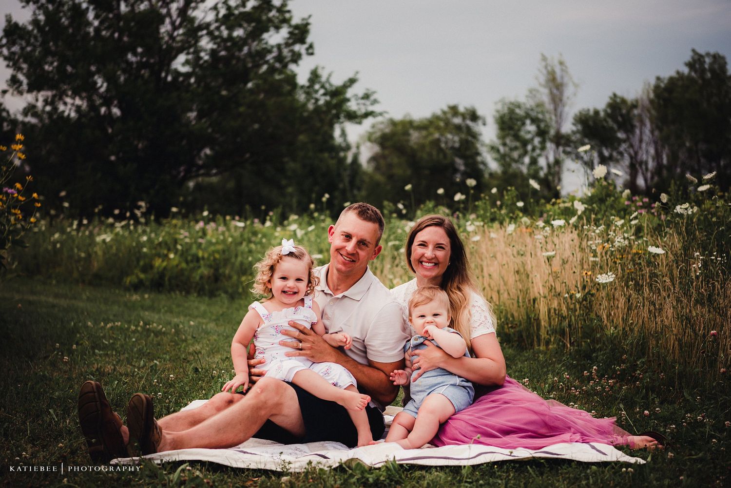 A family sitting together on a picnic blanket in a summer meadow with tall grass and flowers.