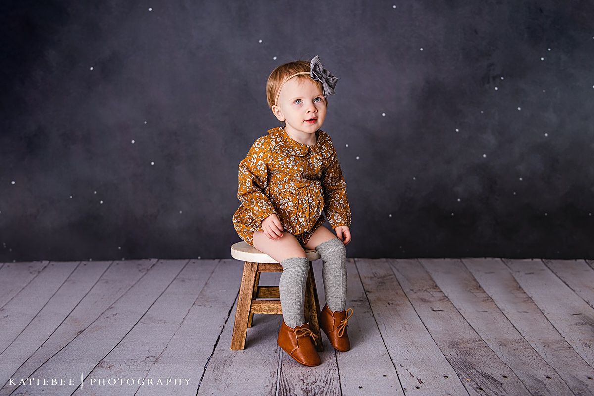 Child in floral pattern outfit sitting on wooden stool against dark sparkly background.