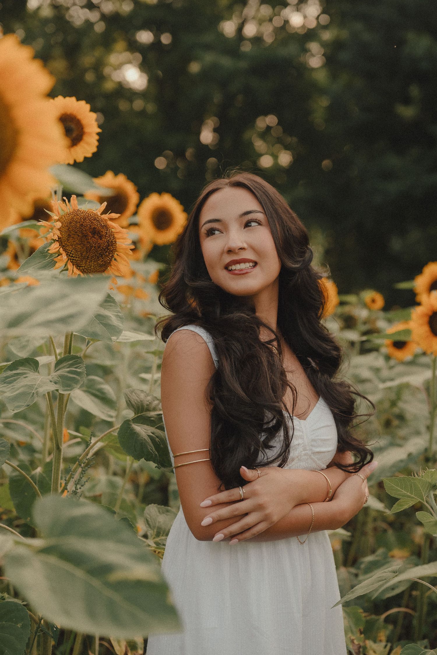 Young woman in white dress smiling among sunflowers in a field, with long dark wavy hair and gold bracelets.