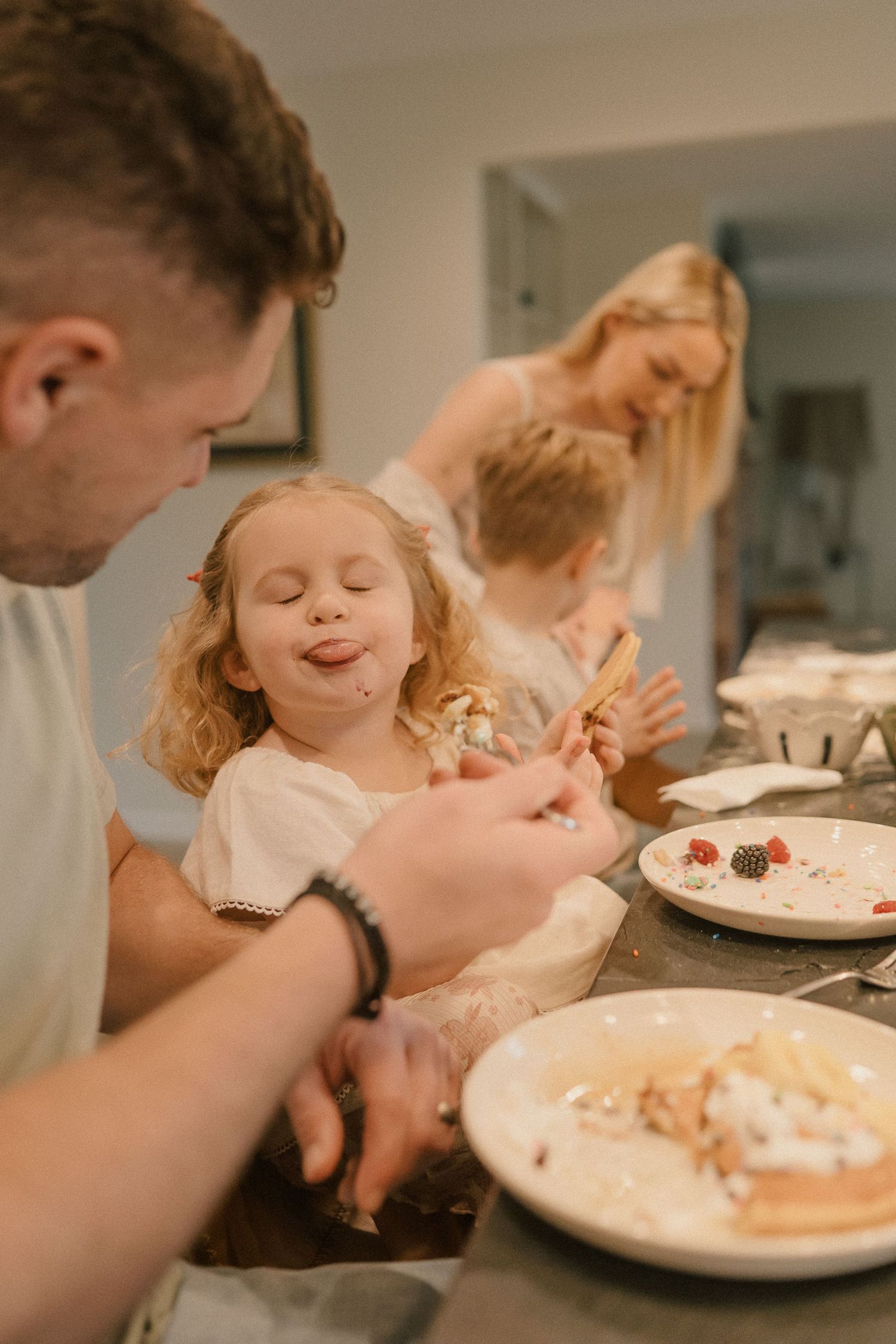 Family with young children decorating pancakes together at a kitchen counter in a bright home.