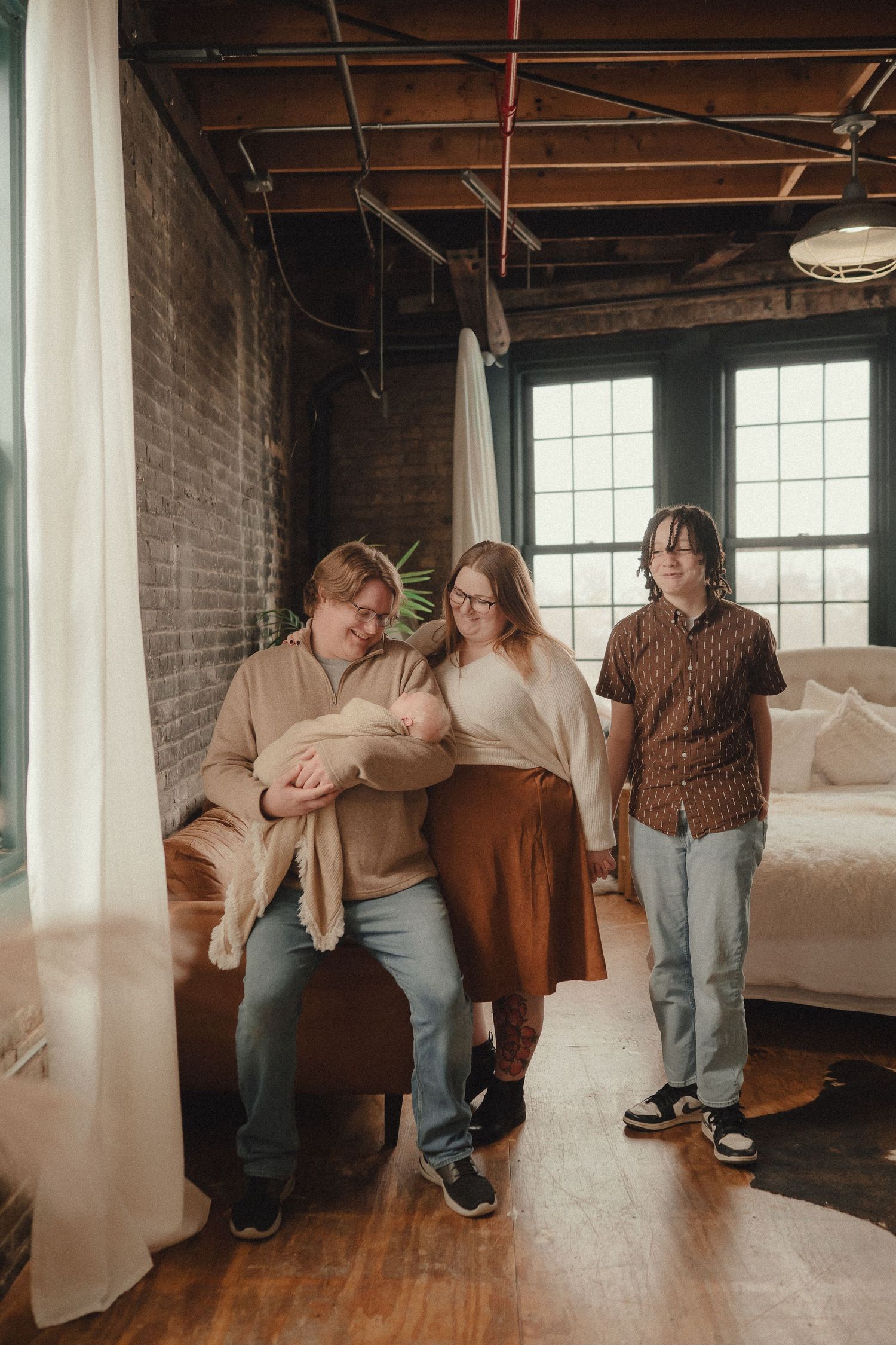 Family of three with newborn baby poses in a cozy industrial loft with brick walls and large windows.