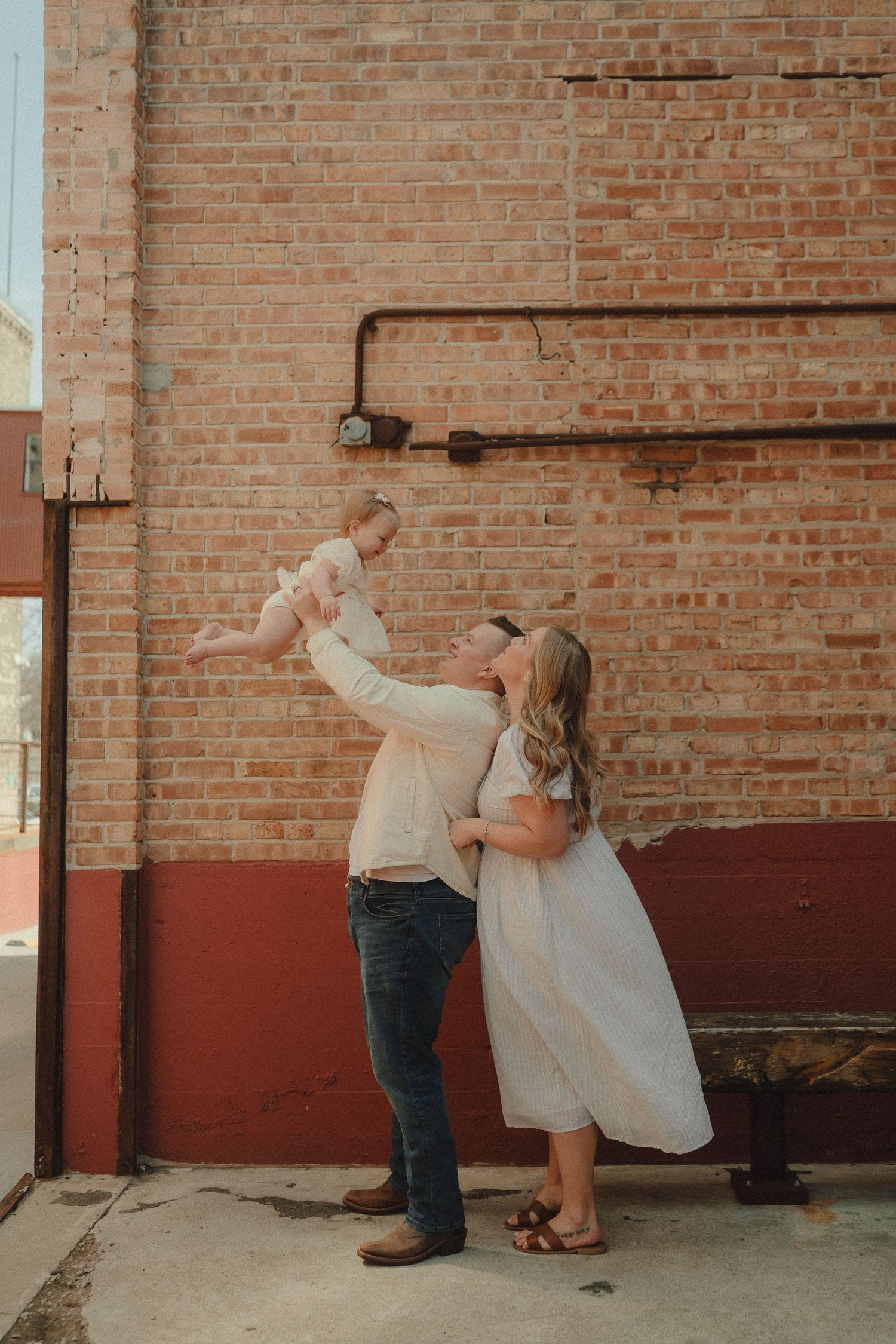 A couple lifts their baby girl in the air together against a warm brick wall during a family photo session.