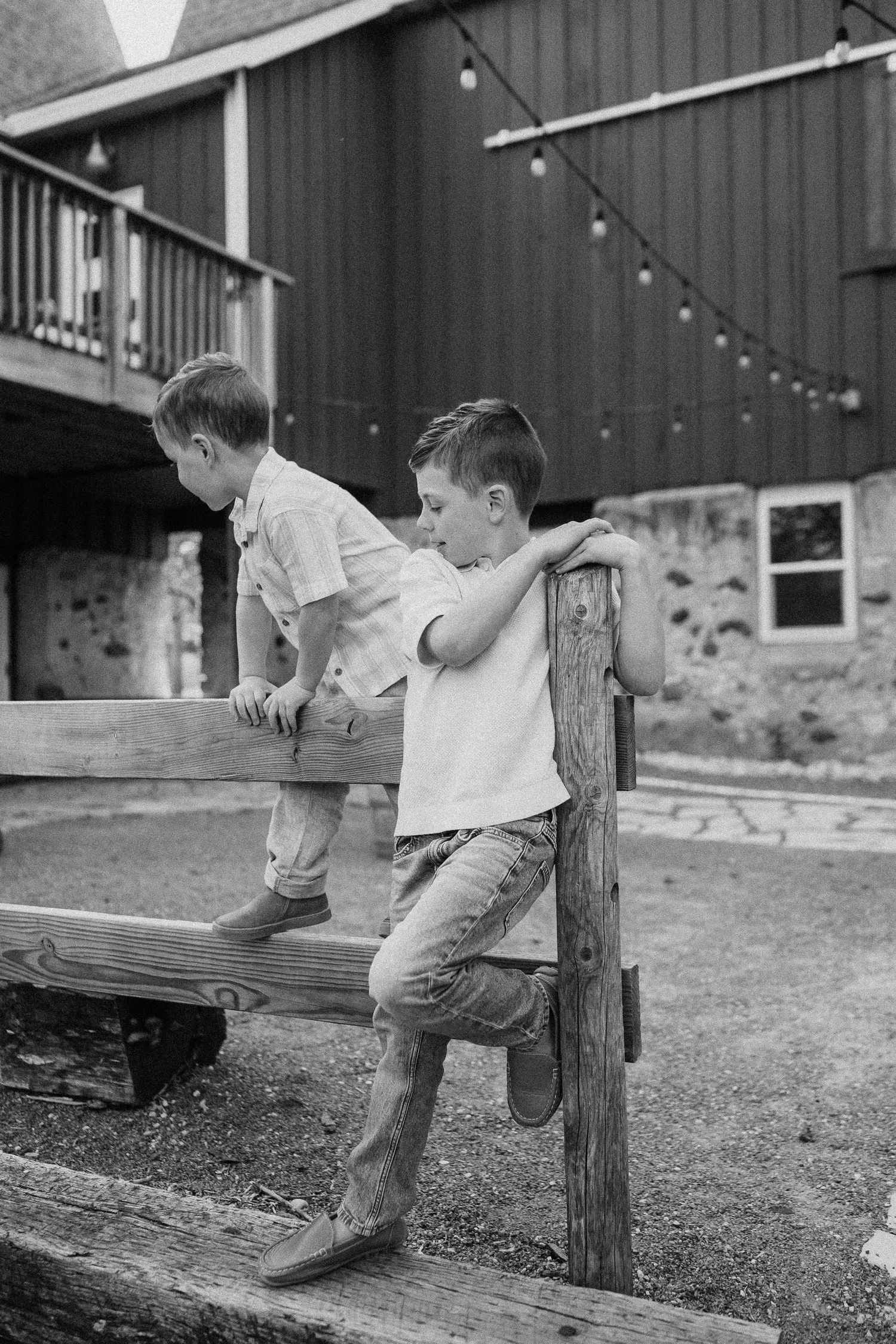 Two young boys playing on a wooden fence near a red barn in black and white photo.