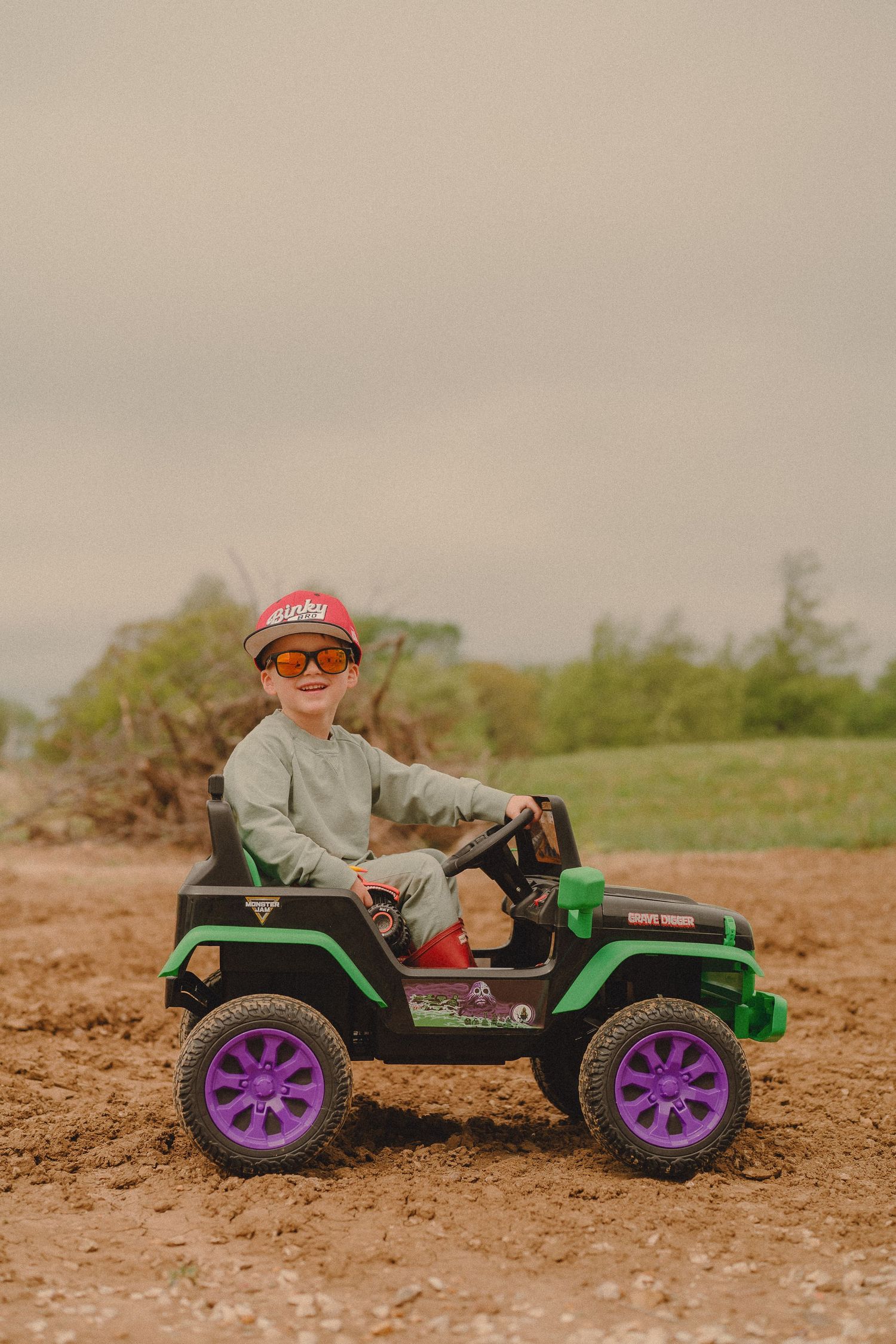 Young boy smiling in Monster Jam toy Jeep on dirt field wearing red cap and sunglasses.