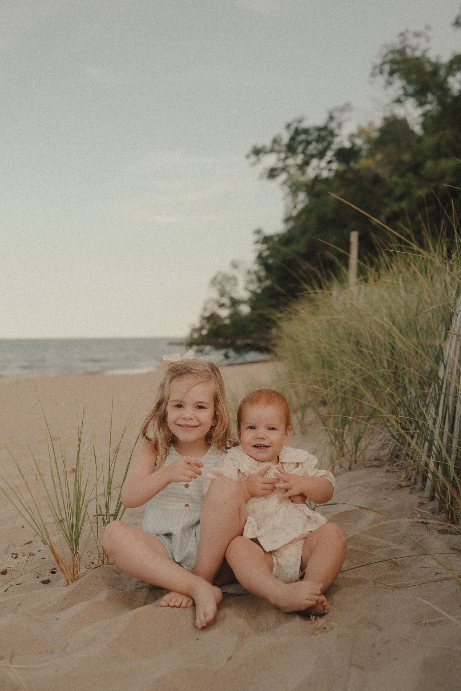 Two young sisters sit together smiling on a sandy beach with dunes and trees behind them.