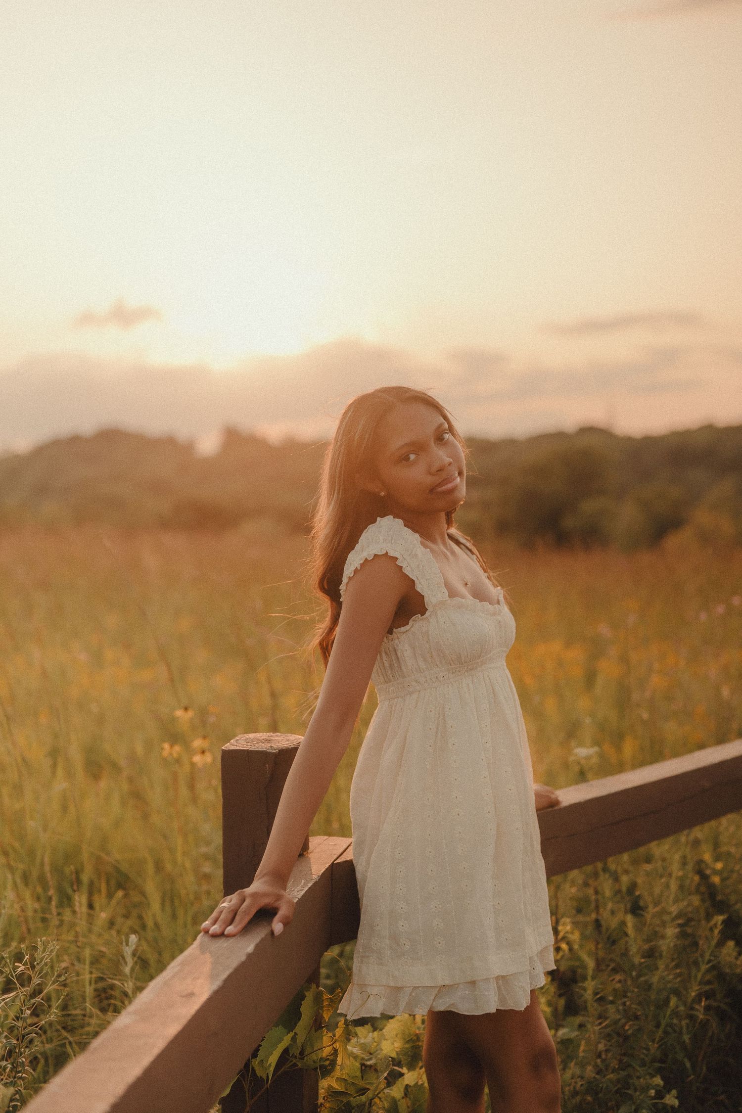 Young woman in white dress leaning on wooden fence in a wildflower field at warm sunset.