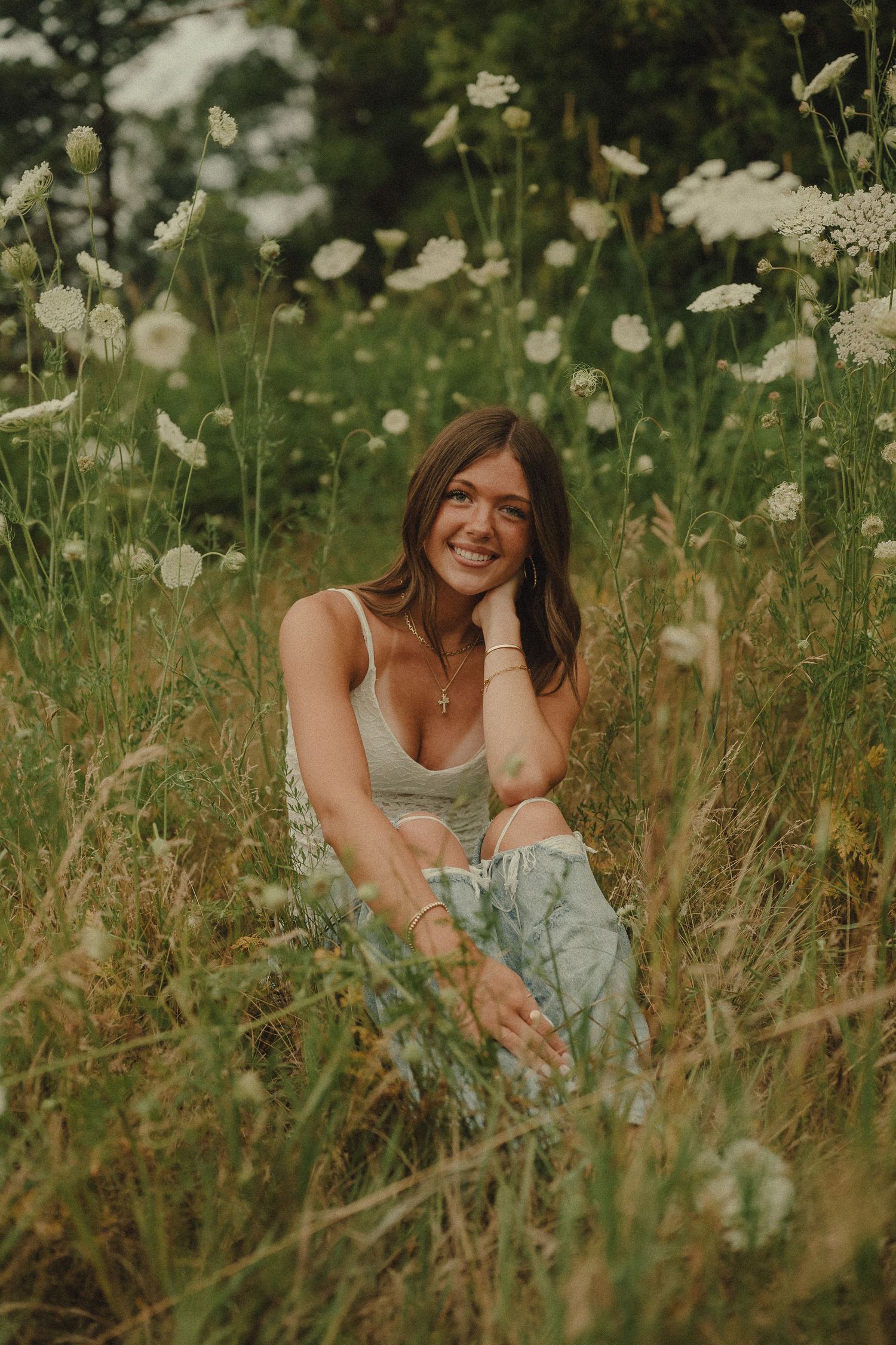 Smiling young woman in white top sitting among tall wildflowers and Queen Annes lace in field.