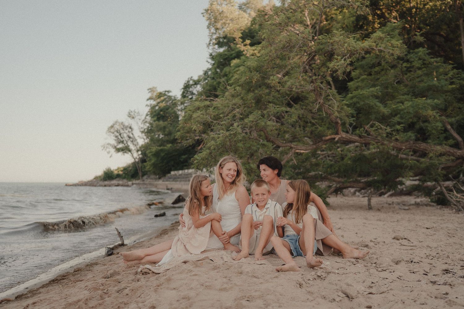 Mother and four children sit closely together laughing on a sandy beach by the water.