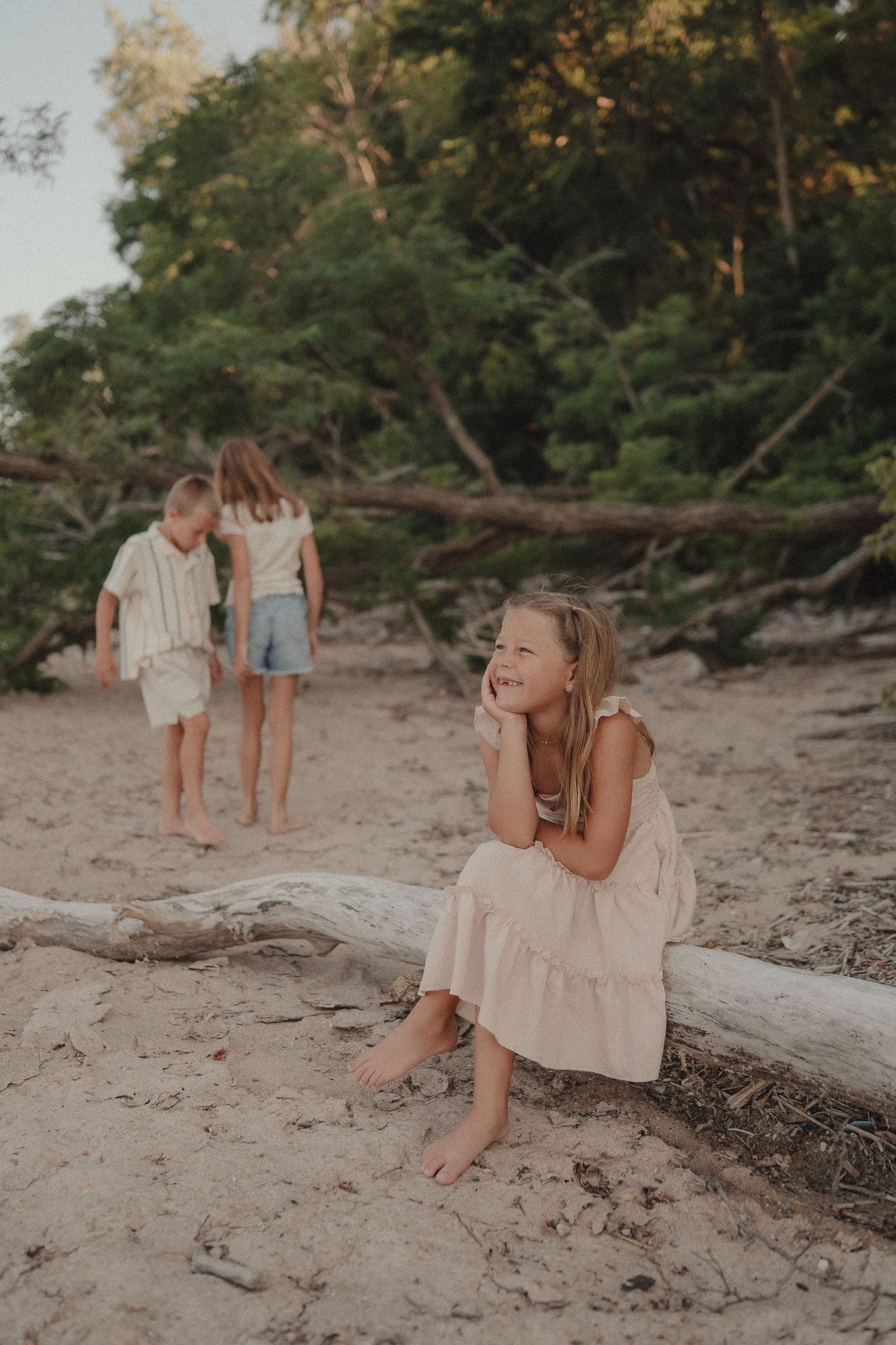 A barefoot girl in a beige dress sits on driftwood smiling on a sandy beach with children playing behind her.