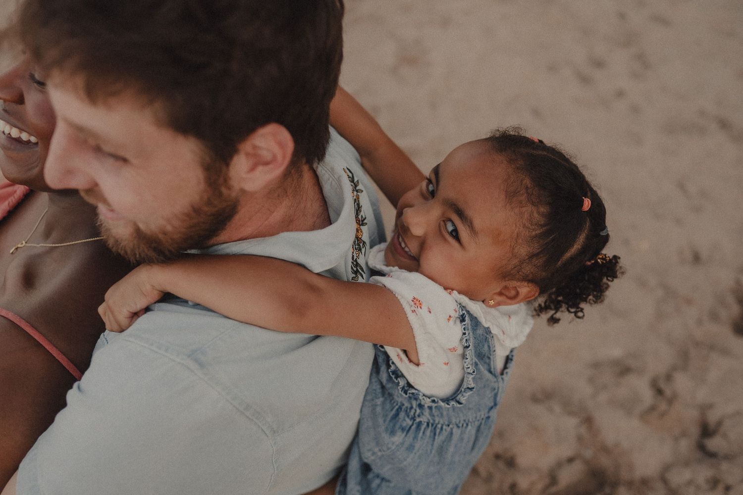 A man gives a joyful young girl a piggyback ride on a sandy beach, both smiling in a candid outdoor moment.