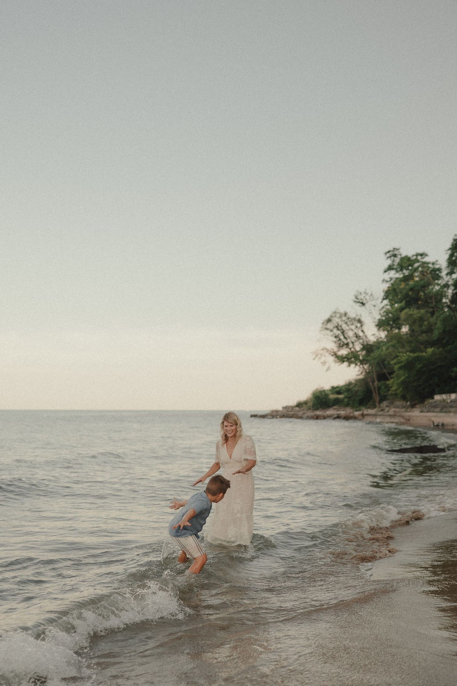 Young boy splashing in gentle ocean waves while his mother in a white dress watches lovingly on a serene beach.
