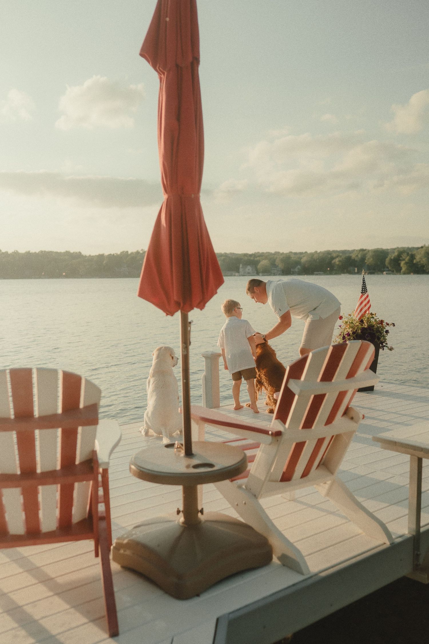 Family enjoying a lakeside dock with Adirondack chairs, red umbrella, and American flag on a sunny day.