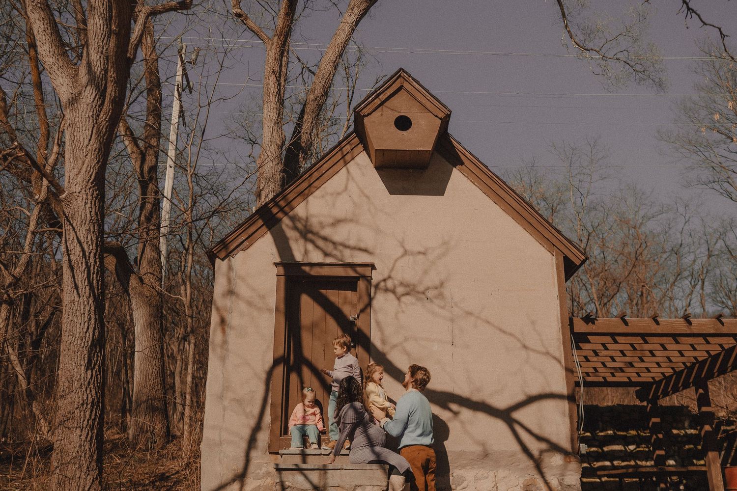 A small beige chapel with a birdhouse on top sits among bare winter trees.