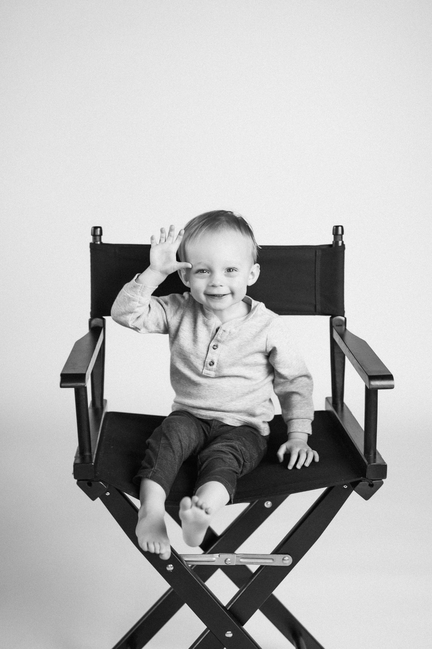 Child waves while sitting in black director's chair during black and white photoshoot.