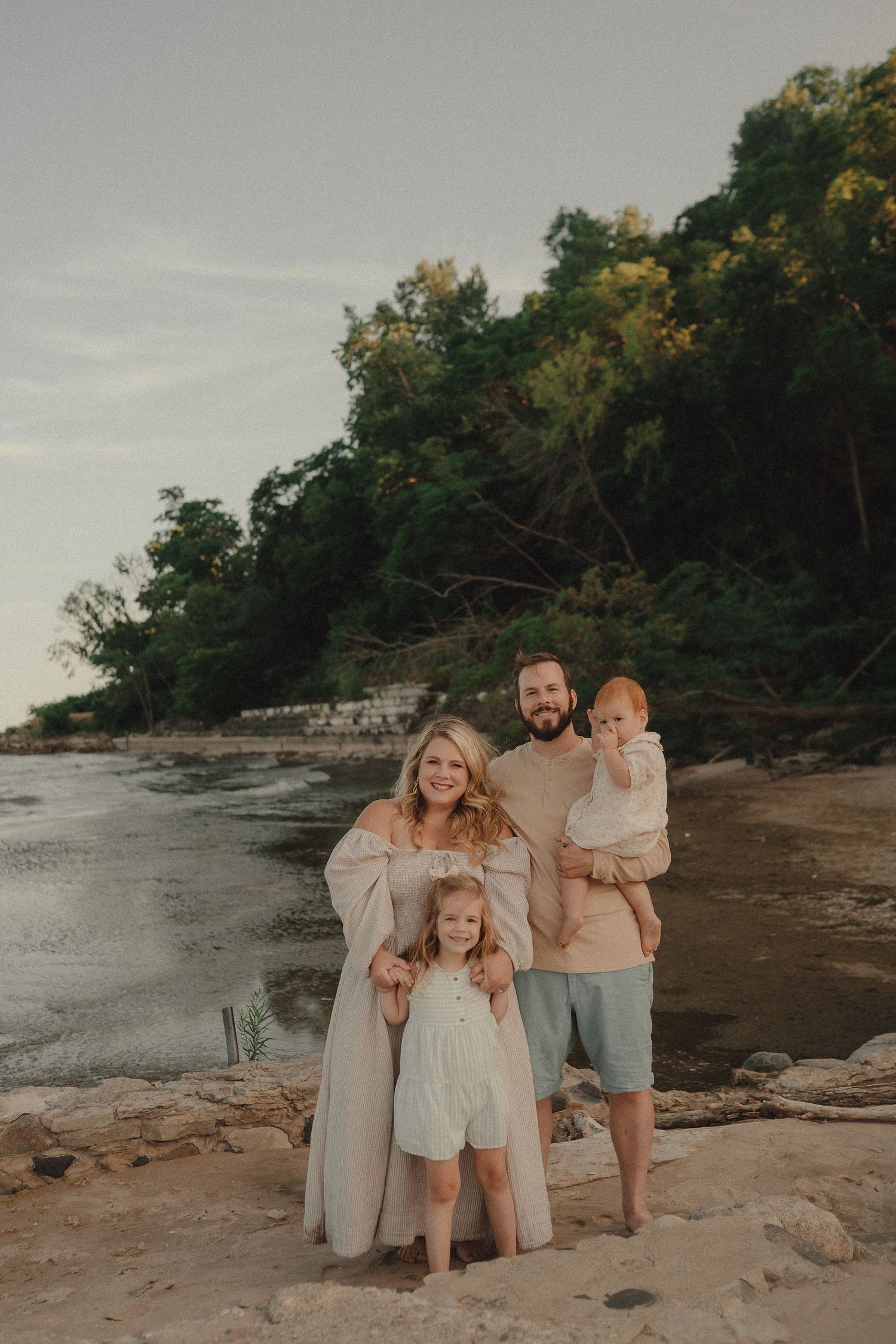 Family of four posing on rocky beach at sunset, parents with toddler and baby near tree-lined shore.