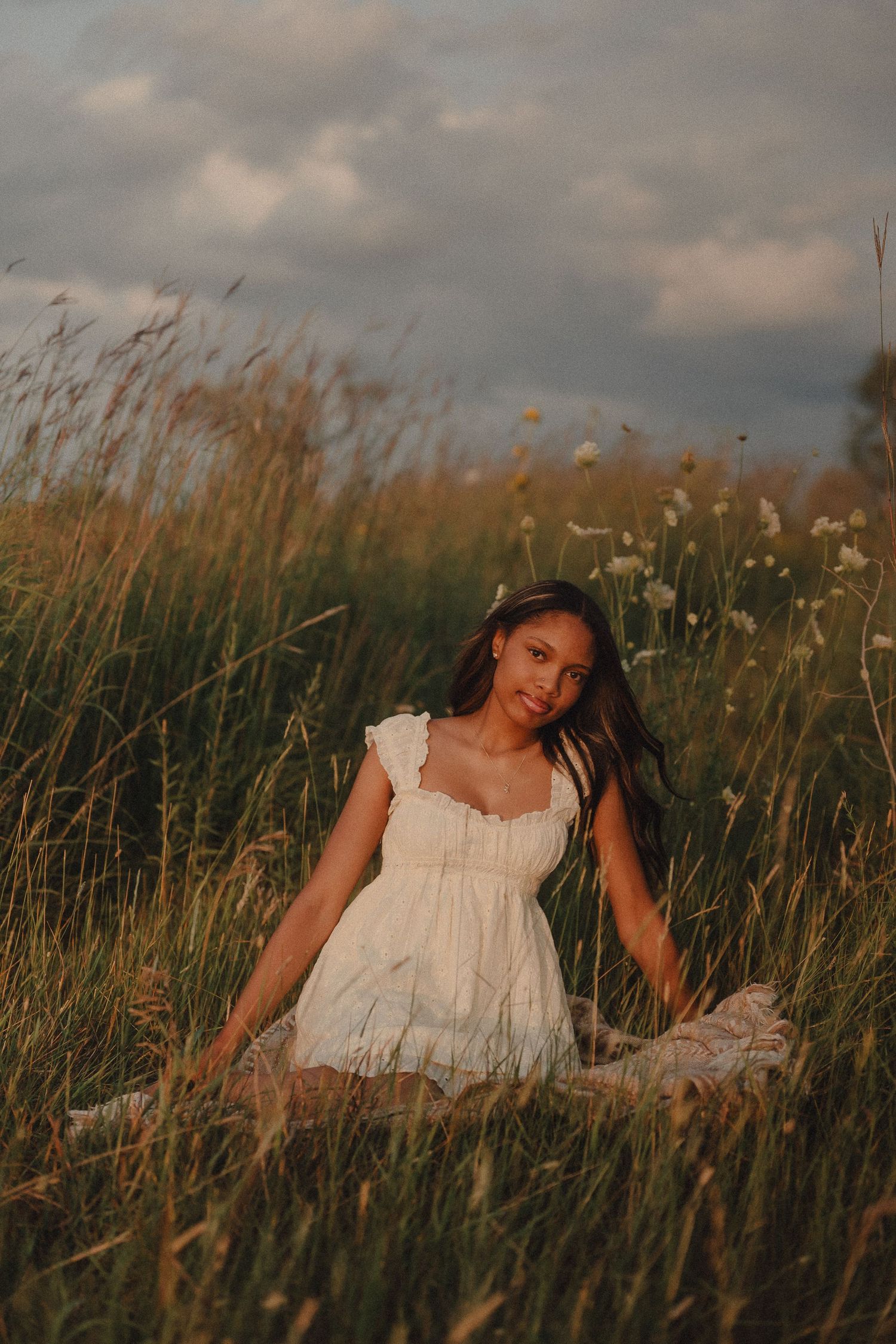 Young woman in white dress sitting in tall golden grass field with wildflowers and dramatic cloudy sky at sunset.