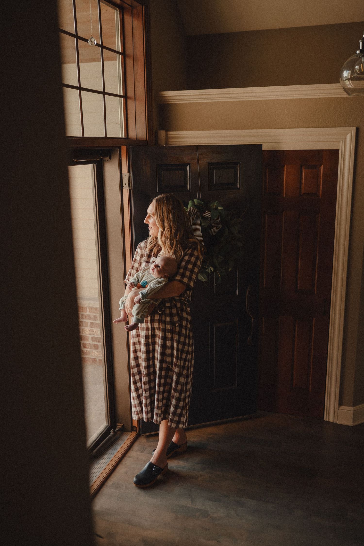 Blonde woman in plaid dress cradling newborn baby while gazing out window in dim interior light.