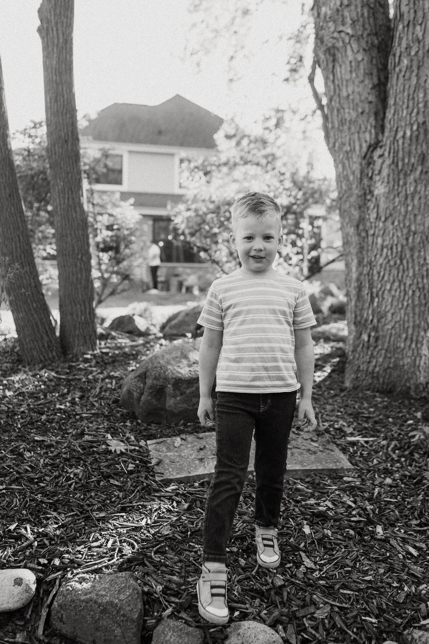 Black and white photo of a smiling young boy in striped shirt and jeans standing in a wooded backyard.